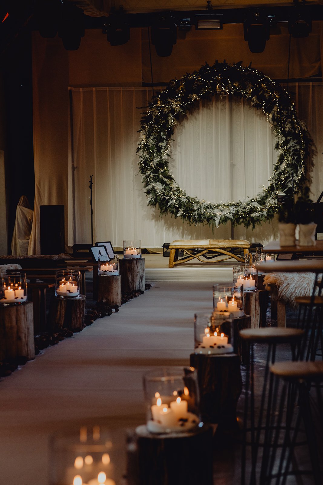 A decorated event space with a large circle wreath adorned with lights and greenery hanging on a white curtain backdrop. Wooden stumps with candles are placed along an aisle, leading to a stage with a wooden bench, creating a cozy and festive atmosphere.