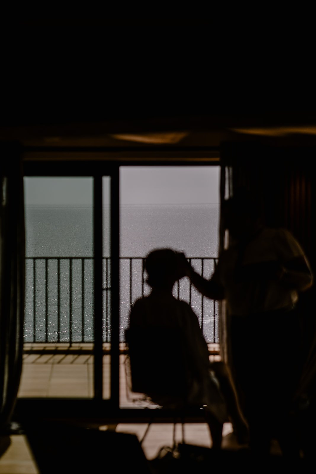 Silhouette of a person receiving makeup or hair styling on an outdoor balcony overlooking the ocean at sunset.