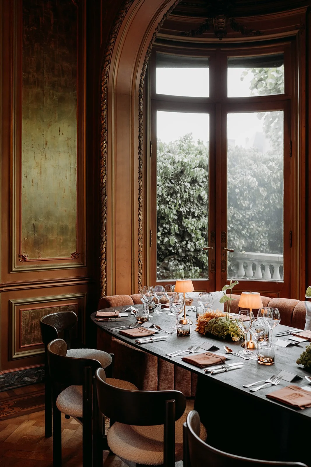Elegant dining room with large arched window overlooking lush greenery, vintage wooden panel walls, a long table set with wine glasses, silverware, napkins, small table lamps, and floral centerpieces, surrounded by vintage-style chairs.