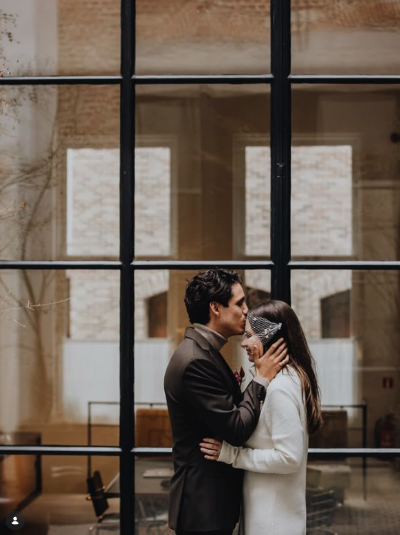 A couple in wedding attire sharing an affectionate moment behind a glass wall, with the groom gently touching the bride's face.