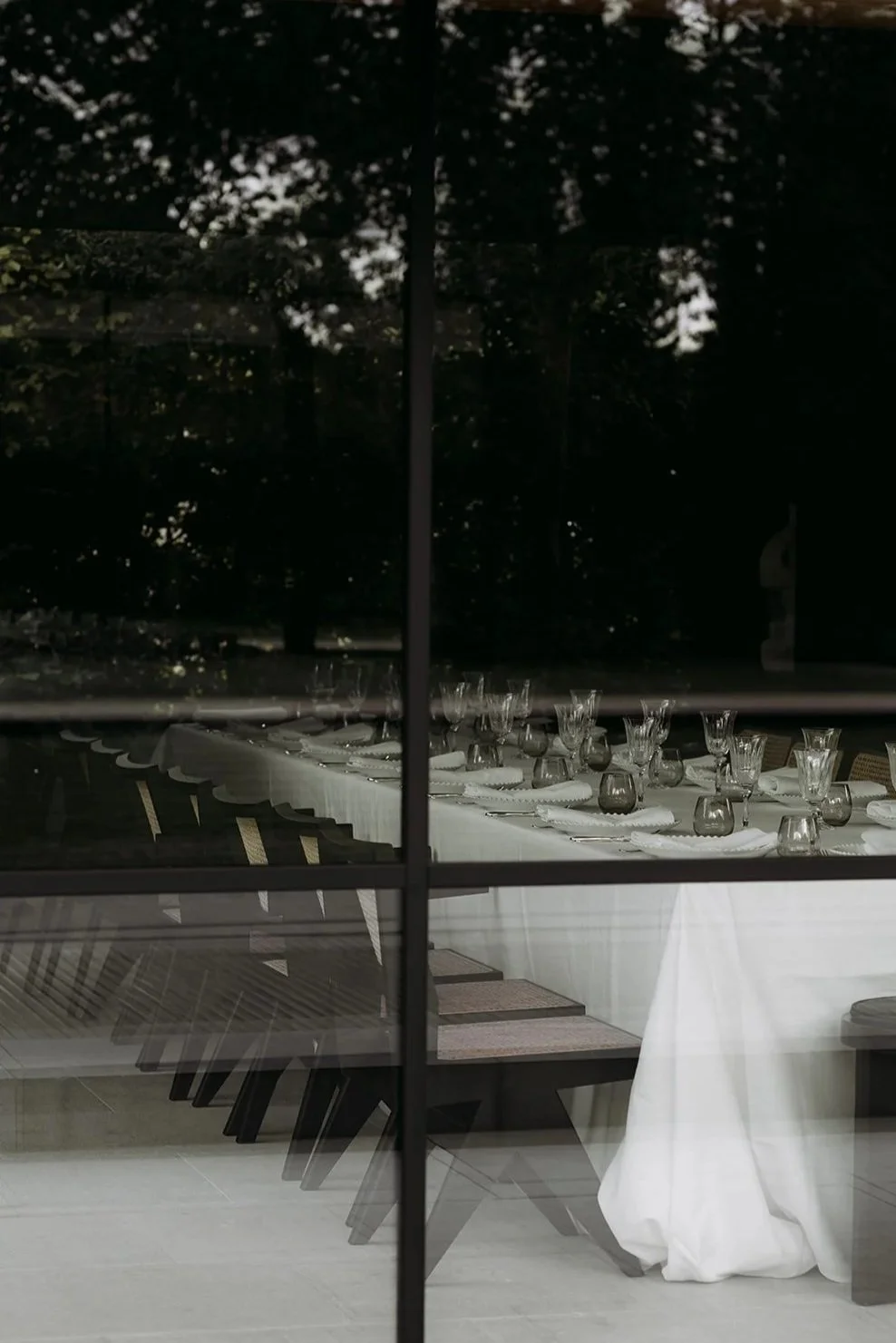 Empty dining table set with white tablecloth, glassware, and plates, seen through a glass window at night, with dark trees visible outside.