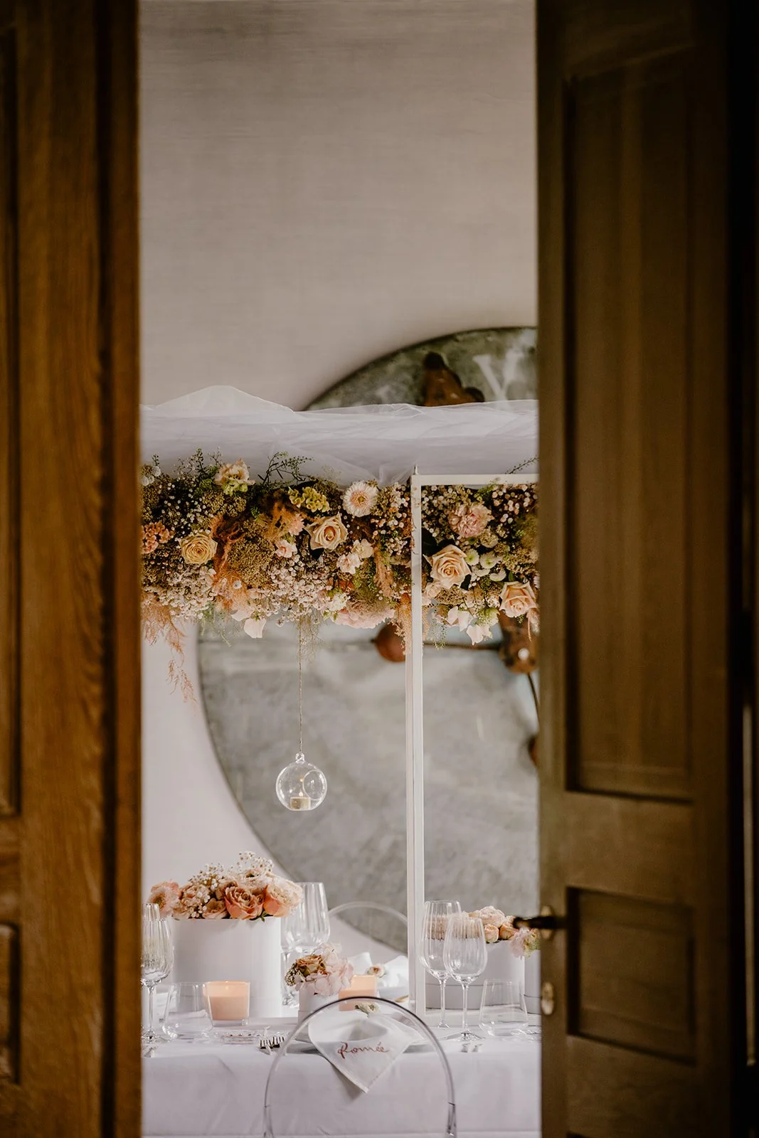 A decorated wedding table with a floral arrangement and candles, seen through partially open wooden doors with a large round mirror in the background.