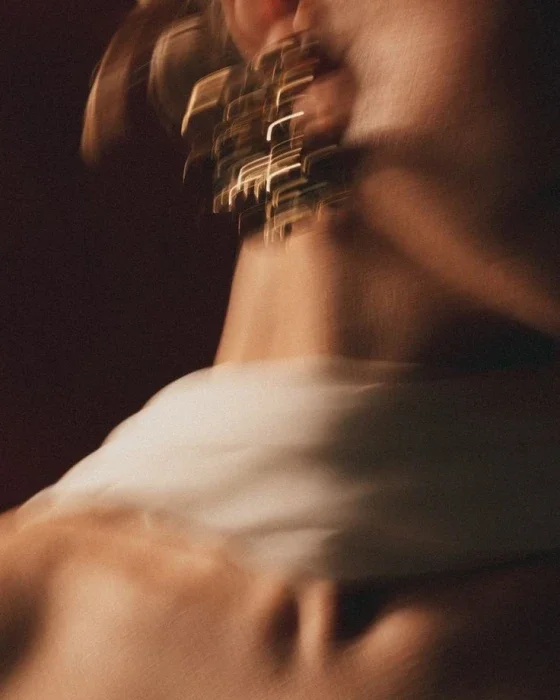 A person wearing a white shirt and large earrings, with their head turned to the side, in a blurry motion shot with warm lighting.