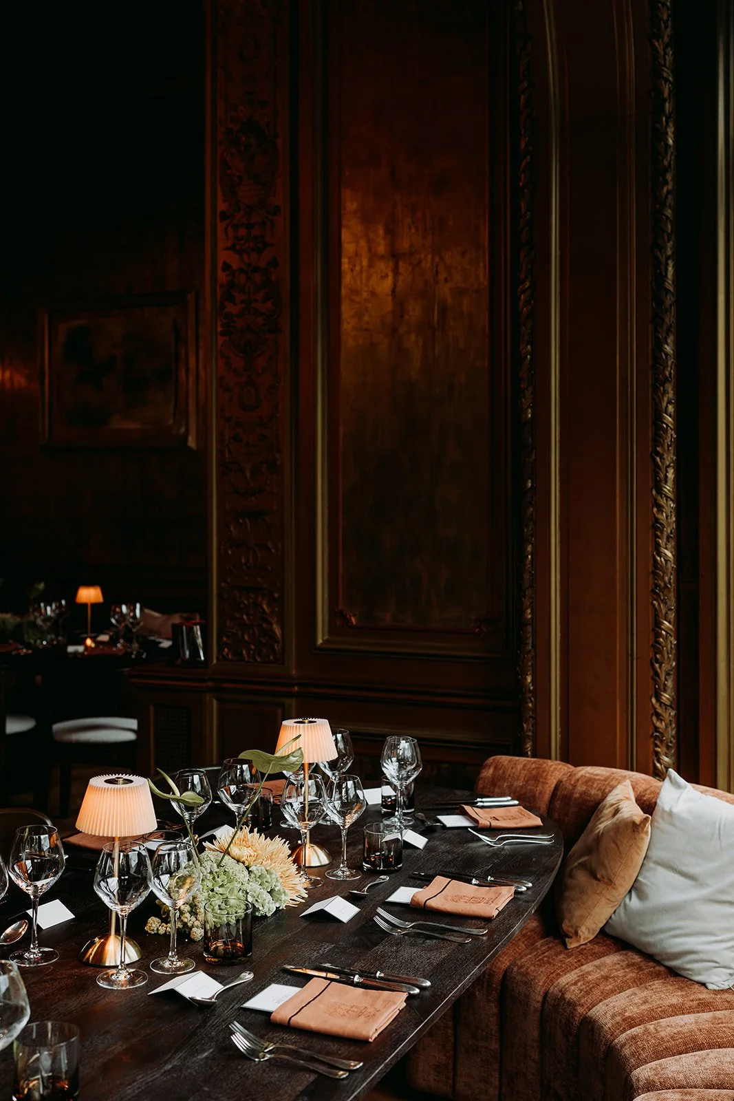 A formal dining room with a dark wooden table set for a meal, decorated with a centerpiece of flowers and flanked by a cushioned banquette and chairs. The table has wine glasses, plates with napkins, cutlery, and small lamps. The background features wood-paneled walls with ornate carvings.