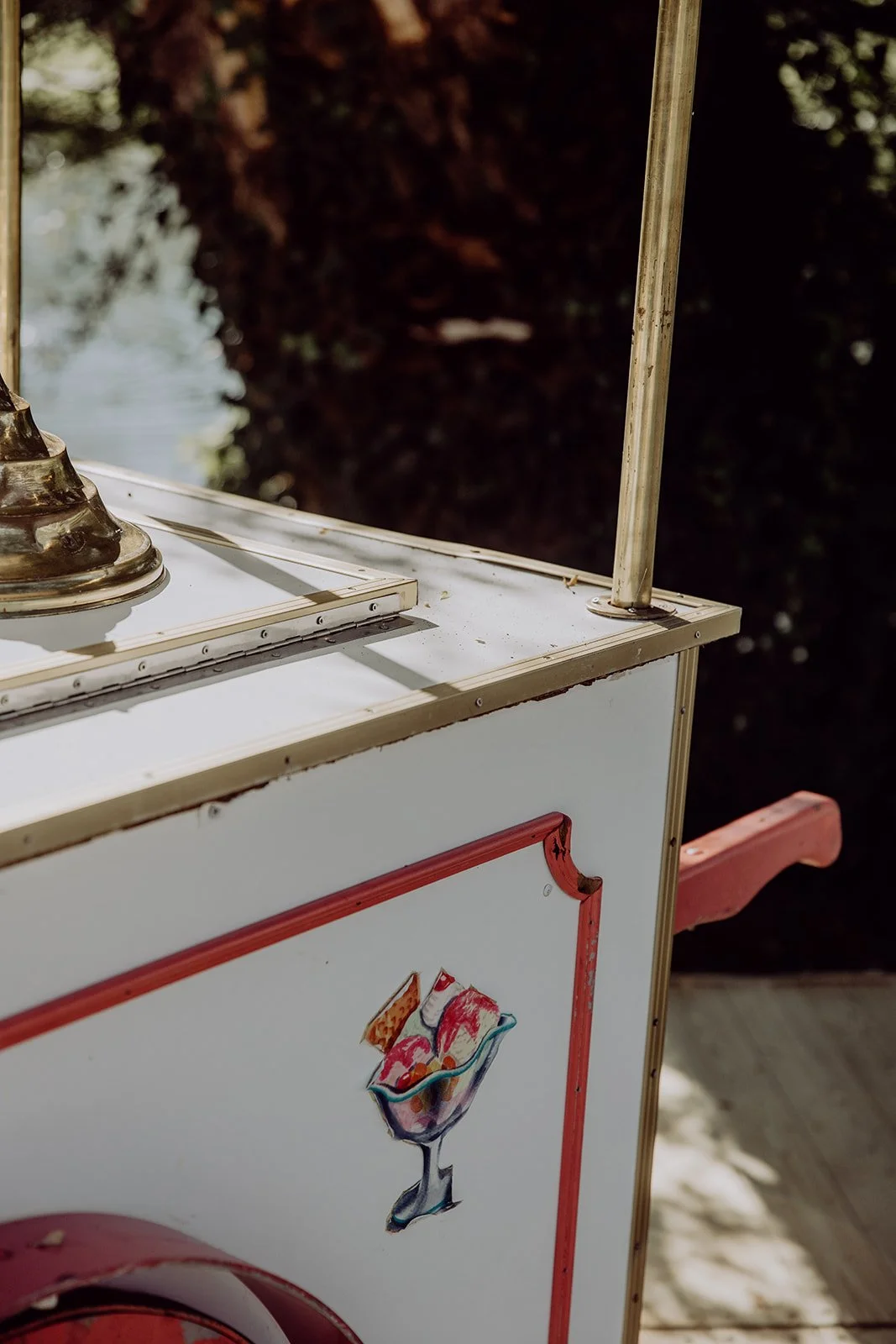 Close-up of a vintage ice cream cart with a drawing of a colorful ice cream sundae on its side, outdoor setting with trees in the background.