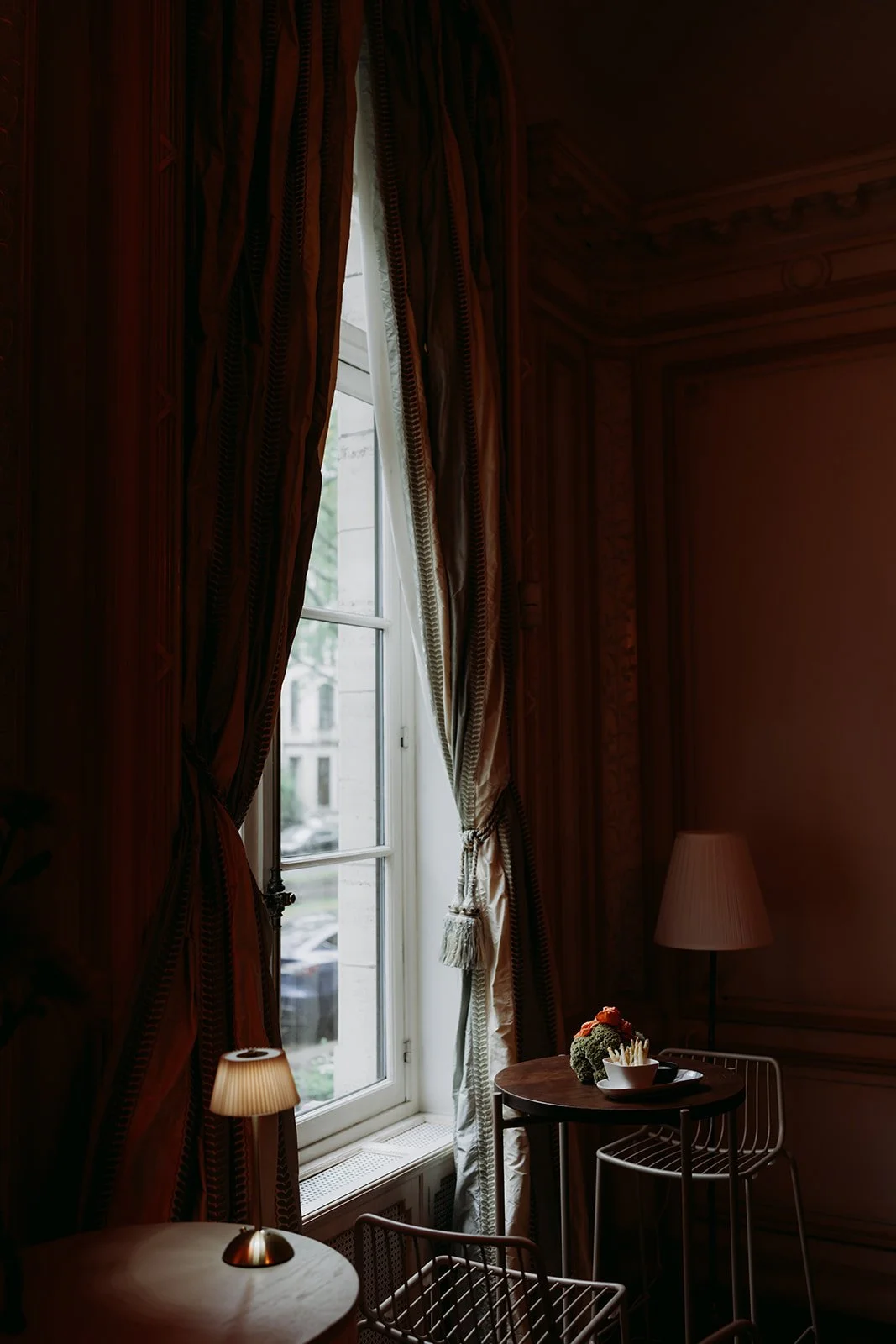 An interior scene with a tall window framed by ornate patterned curtains, next to a small round table with a bowl of food and decorative items, along with a modern wire chair, in a dimly lit room with warm lighting.