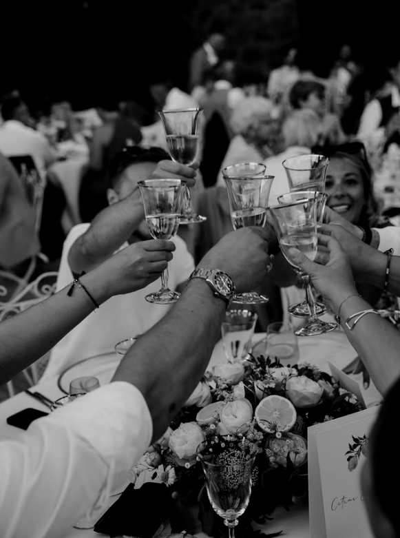People raising champagne glasses in a toast at a celebration or wedding, with a table adorned with flowers and a group of smiling guests in the background.