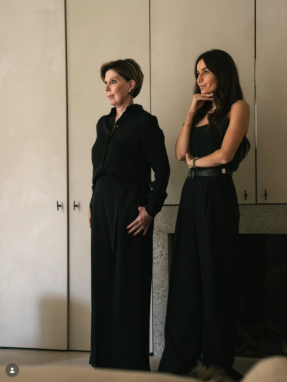 Two women standing indoors, both dressed in black, facing slightly to the side. The older woman has short hair and is wearing a long-sleeve black blouse and wide-leg pants. The younger woman has long dark hair, is wearing a sleeveless black top and high-waisted black pants, and is resting her chin on her hand with a thoughtful expression.