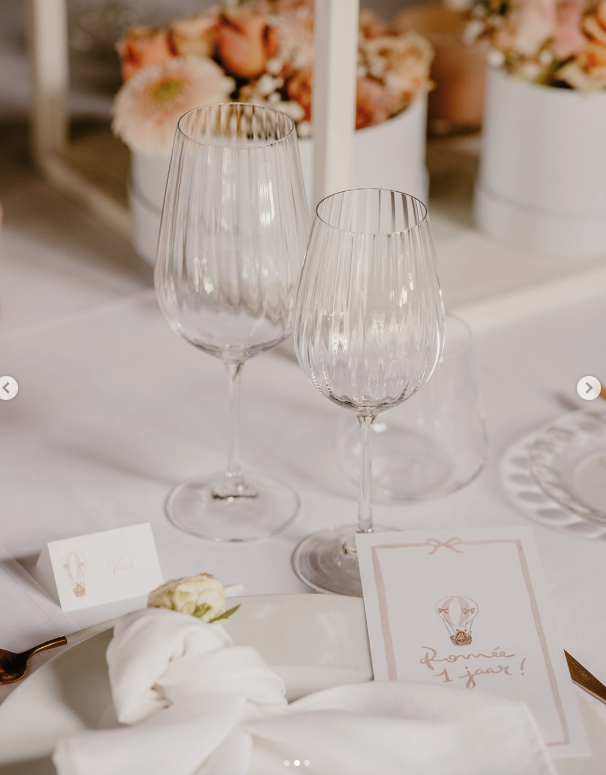Two empty clear wine glasses on a table with floral decorations in the background and a pink and white card on the table.