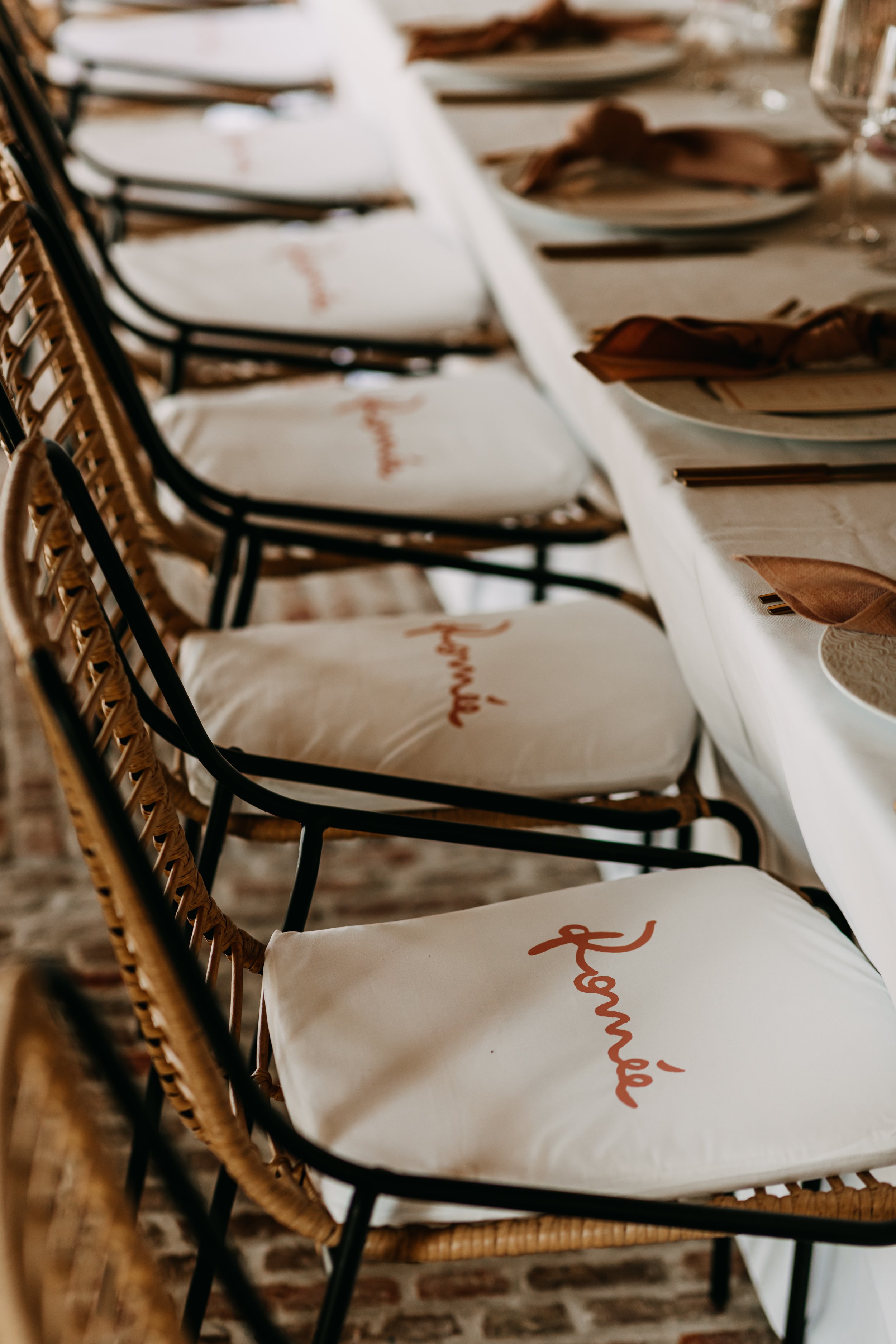 A row of chairs with wicker backs and white cushions with orange cursive writing, set at a dining table with plates, napkins, and silverware in a restaurant or cafe setting.