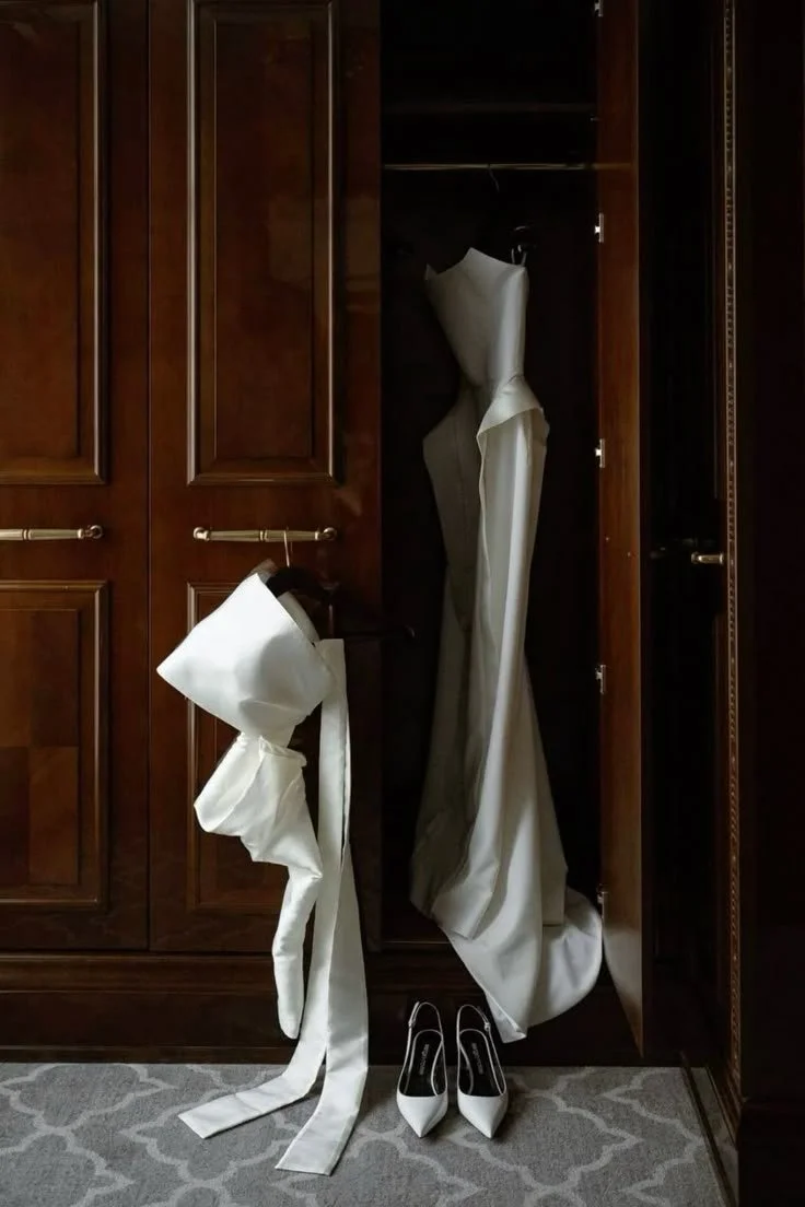 A wooden closet with open doors showing white wedding dress and veil. A pair of white high-heeled shoes placed on the carpeted floor.