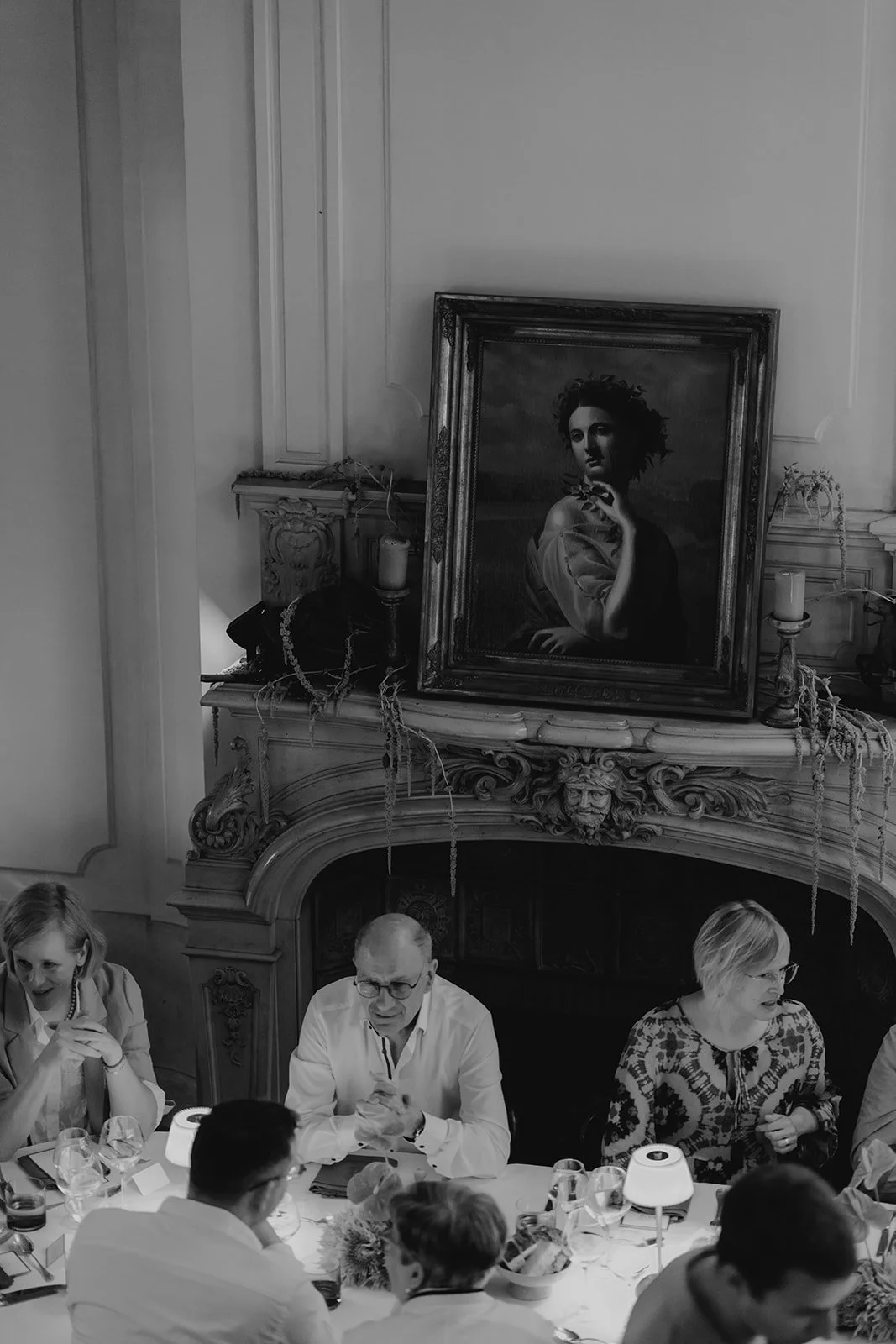 People sitting at a dining table in an elegant room with ornate fireplace and large portrait painting above it. The photo is in black and white.