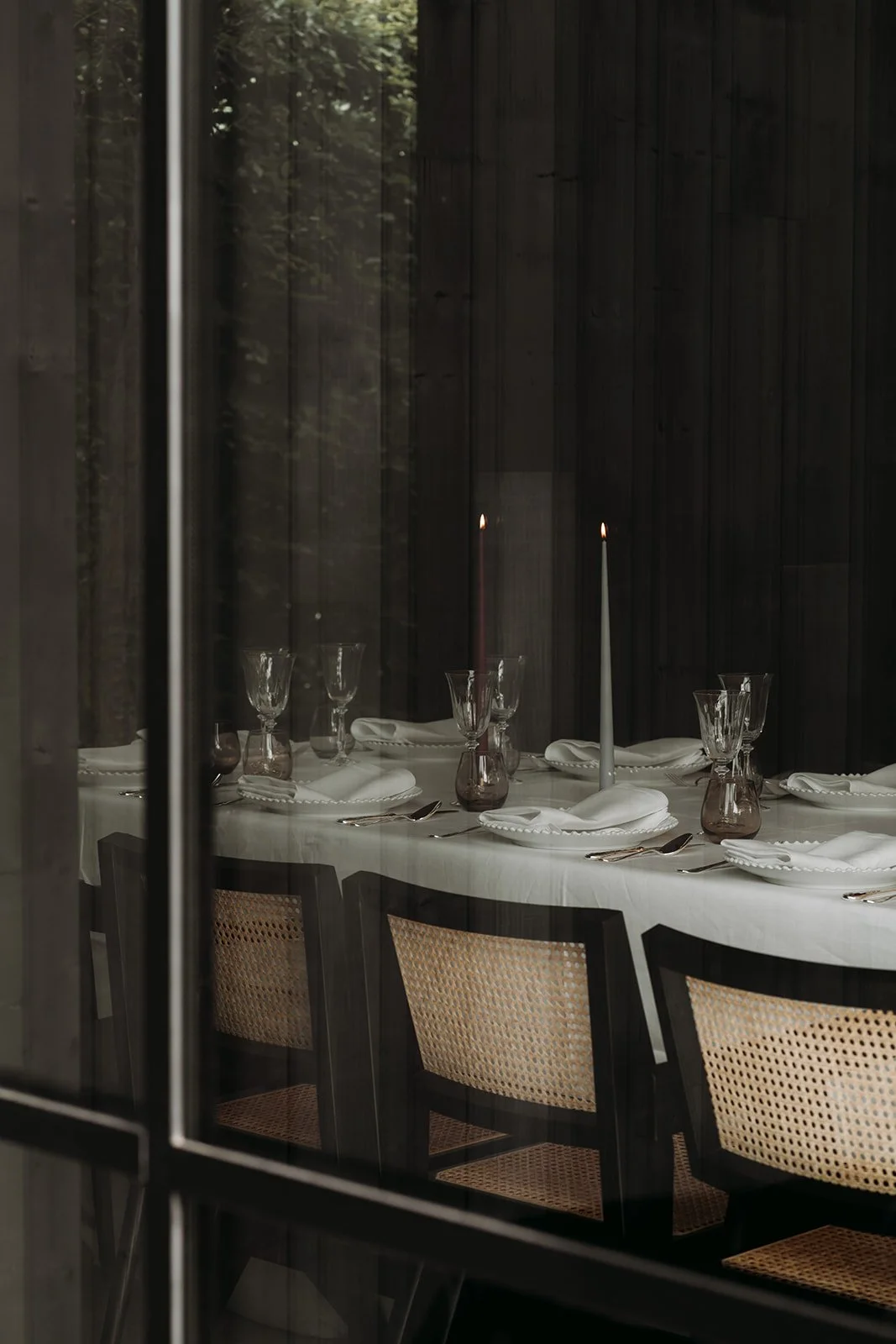 A dining table set with white cloth, white napkins, wine glasses, and two lit candles inside a dark room, viewed through a window.