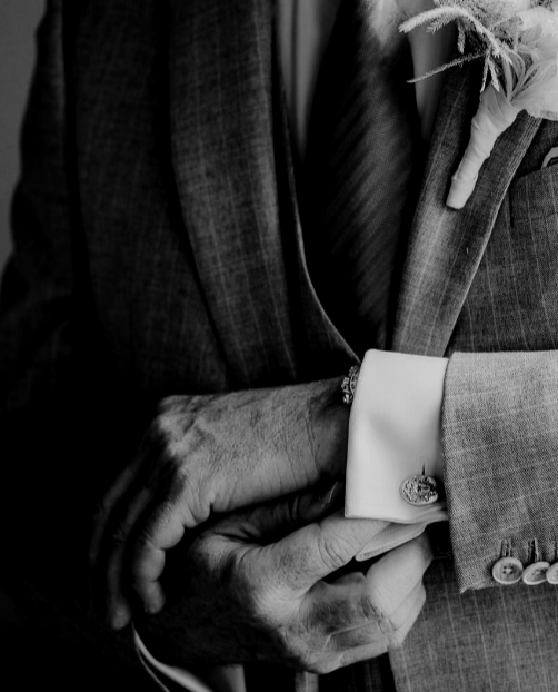 Close-up of a person's hand with a wedding ring, holding a bouquet of flowers, dressed in a suit with cufflinks, in a black and white photo.