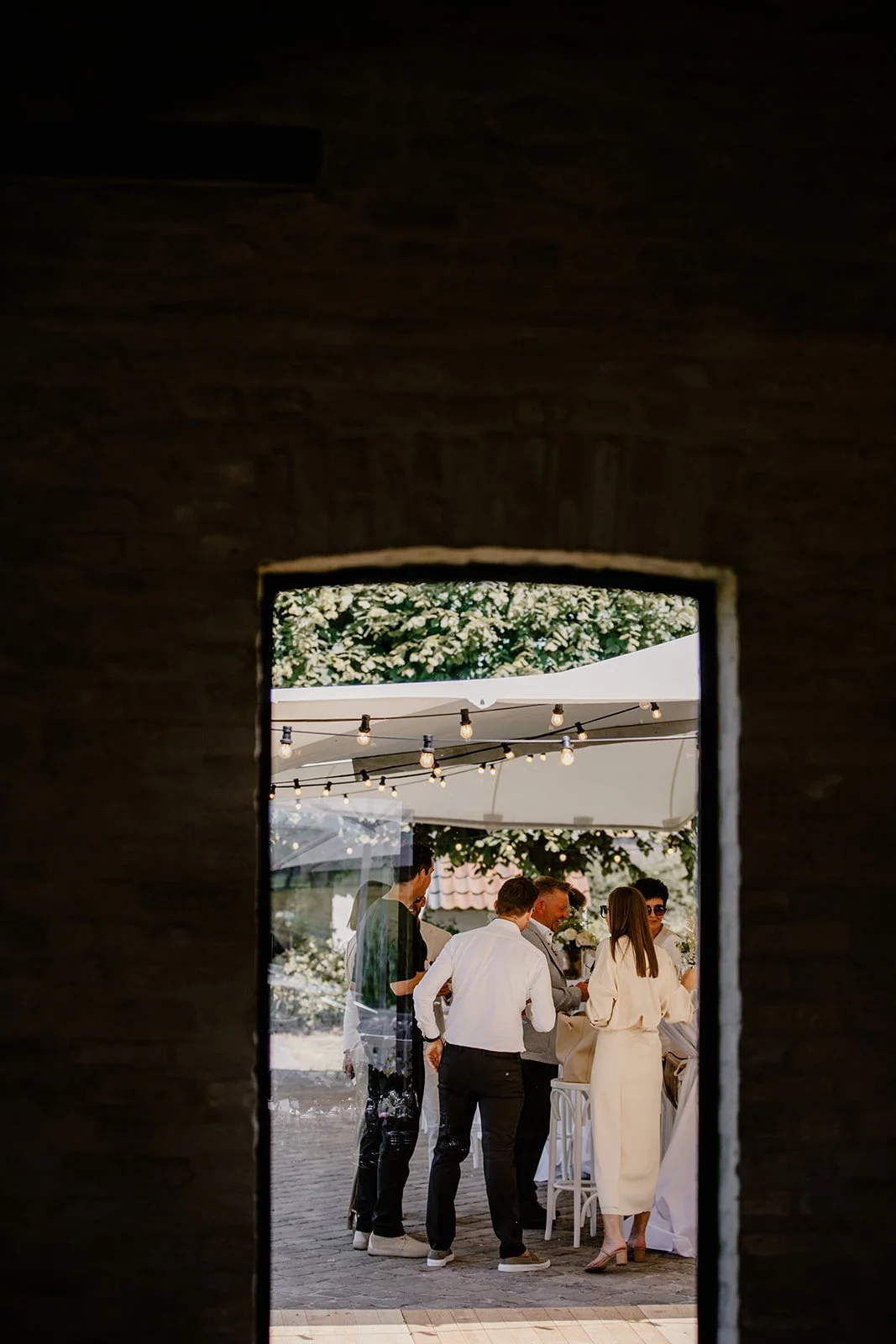 People gathered at an outdoor event, seen through a small window in a brick wall. The setting includes string lights and a canopy, with individuals engaged in conversation.
