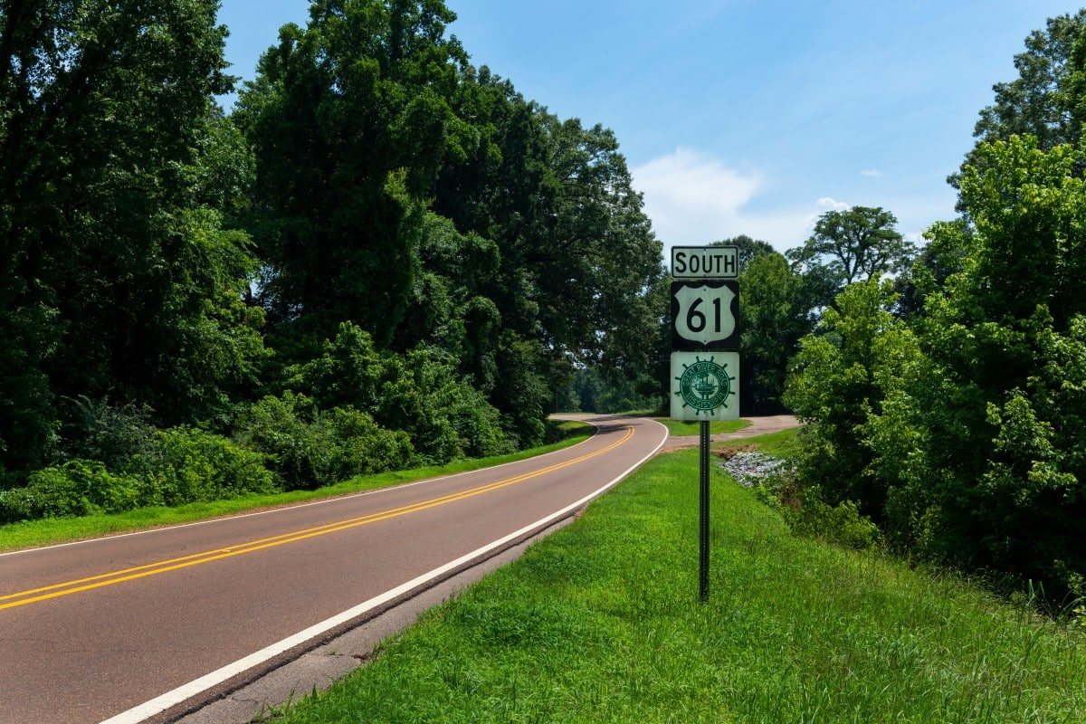 A winding two-lane road surrounded by green trees and bushes on both sides, with a sign indicating southbound US Highway 61.