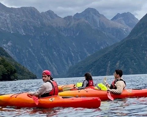 Three people in kayaks in fiords of New Zealand with mountains in the background.