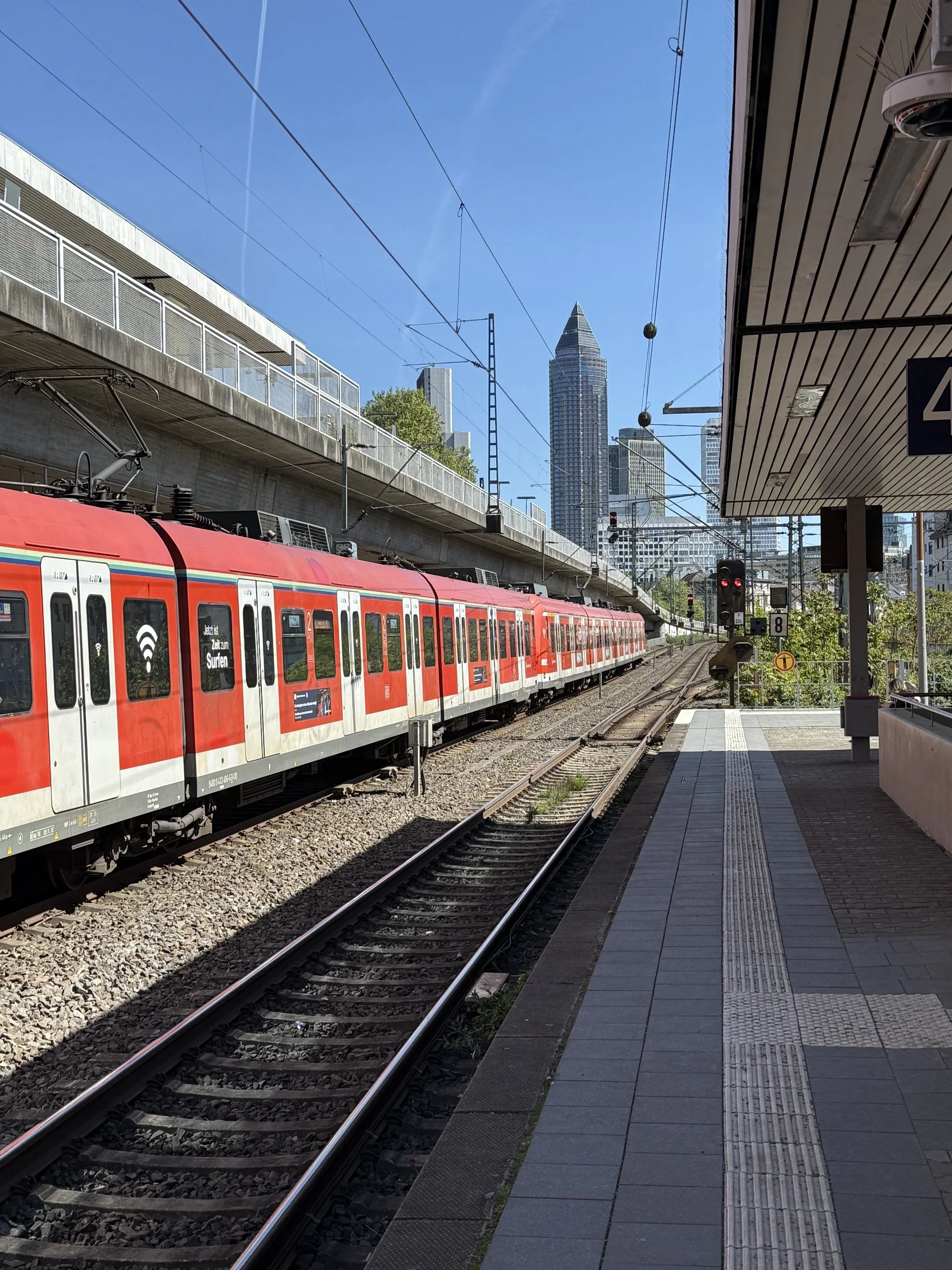 A red train at a train station in Frankfurt Germany with a blue sky and tall skyscrapers in the background. The platform is empty with a patterned surface, and there are overhead electrical lines above the train.