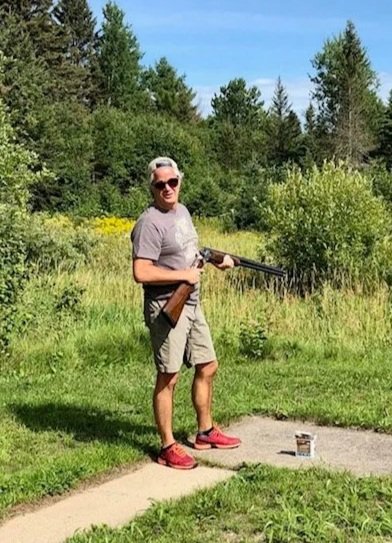 A man standing outdoors on a grassy area holding a shotgun, wearing sunglasses, a gray t-shirt, beige shorts, and red shoes, with a forest background under a blue sky.