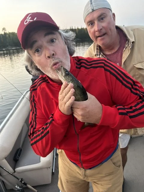 Two men outdoors near a body of water, one holding a fish and making a surprised face, the other standing behind with a neutral expression.