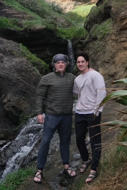 Two people standing in front of a small waterfall in New Zealand in a rocky, outdoor area with green vegetation.