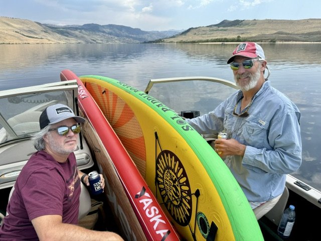 Two men on a boat with inflatable paddleboards, holding drinks, in a lake surrounded by hills.