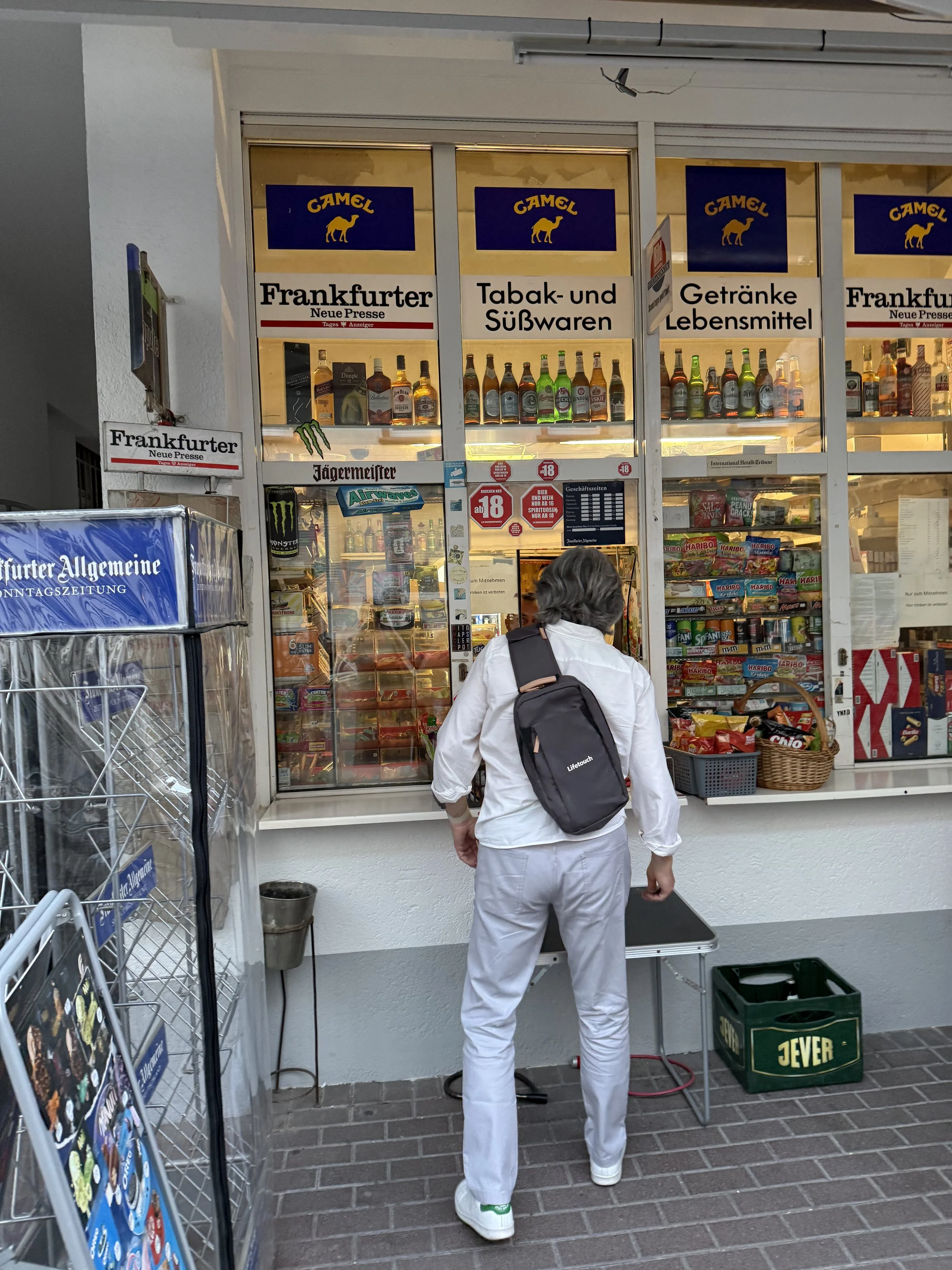 A person dressed in white, carrying a black backpack, standing outside a kiosk shop with snacks and beverages, including alcohol, behind glass windows. The shop has signs in German and bottles of alcohol displayed inside.