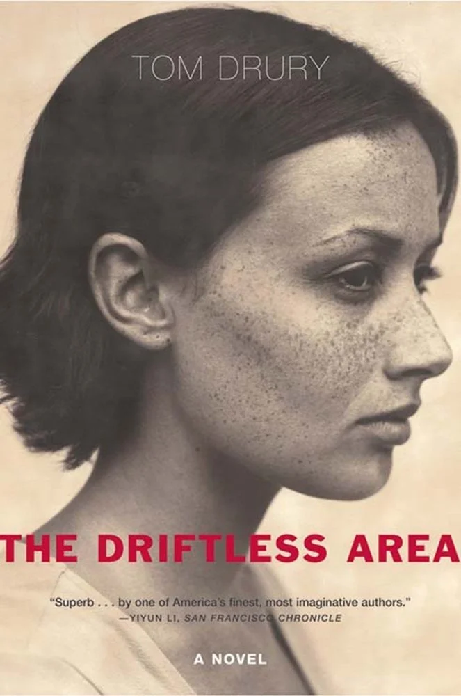 Book cover for 'The Driftless Area' by Tom Drury, featuring a black-and-white profile of a woman with freckles and dark hair.