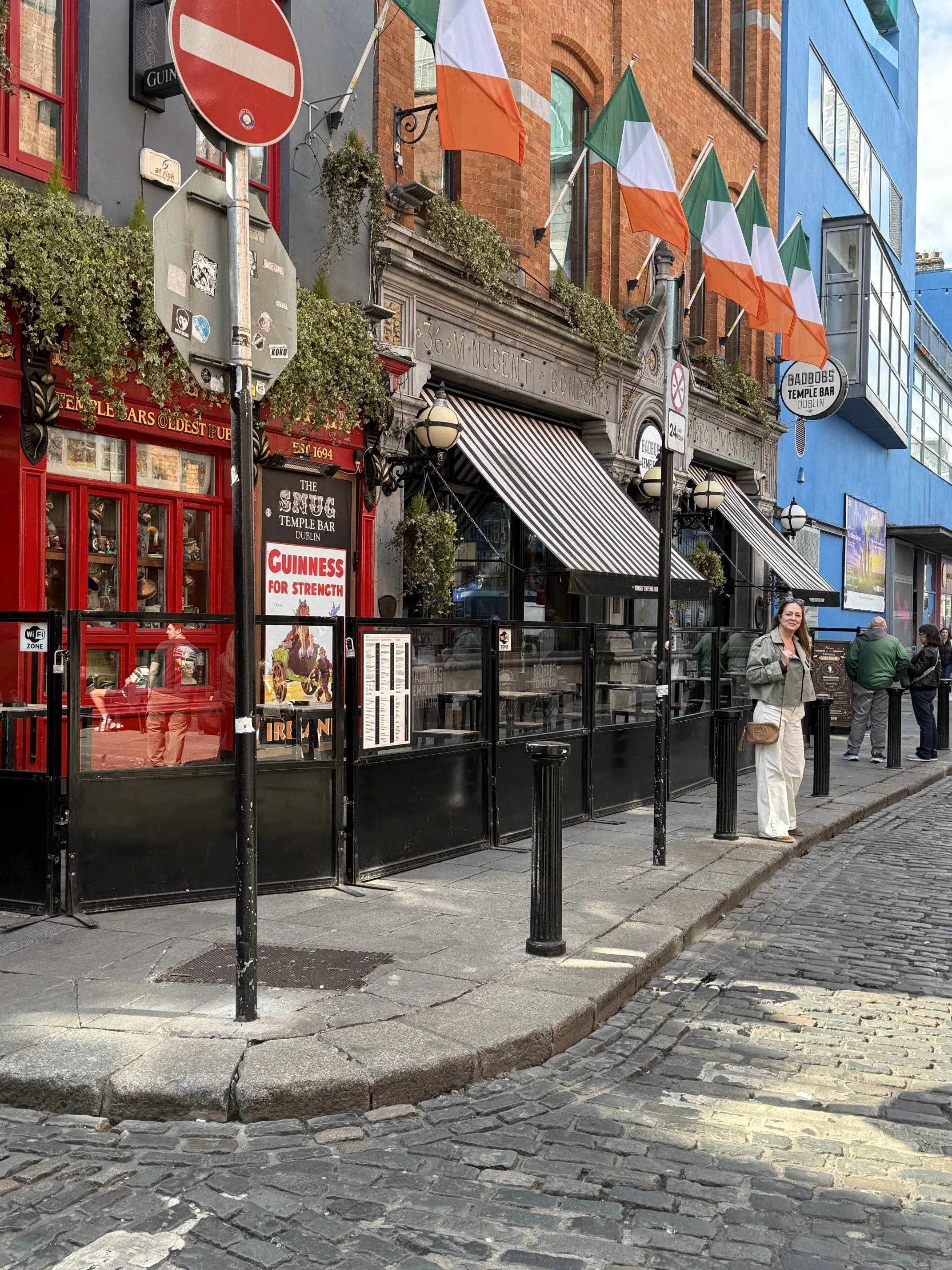 Street view of a bar or pub with Irish flags, outdoor seating, and a woman standing on the sidewalk. The sign indicates it is 'The Snug Temple Bar Dublin.' Red and black accents are visible on the building's exterior, with some greenery and hanging plants.