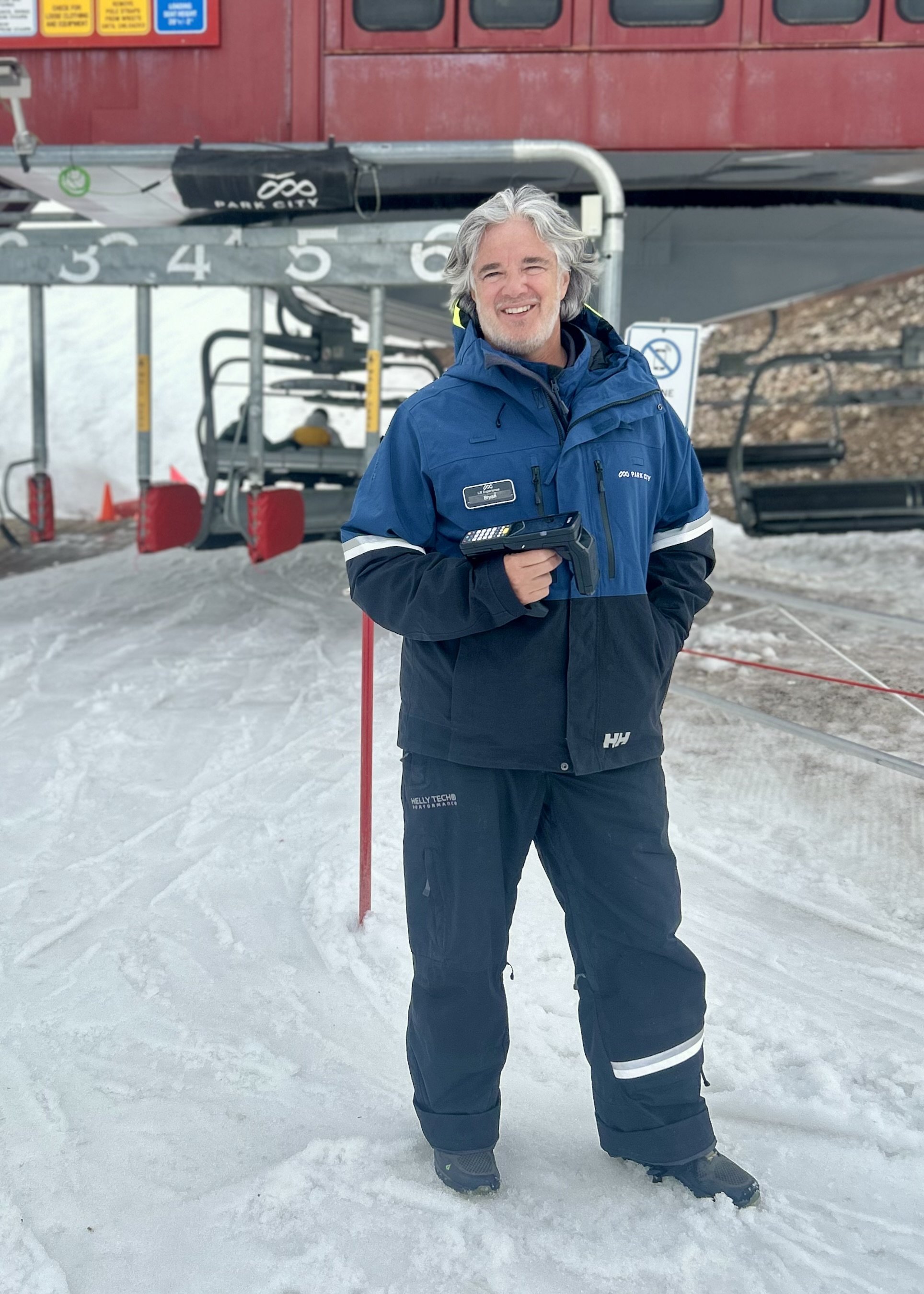 A man smiling while standing on snow at a ski resort, holding a payment terminal, with ski lift equipment and a red building in the background.