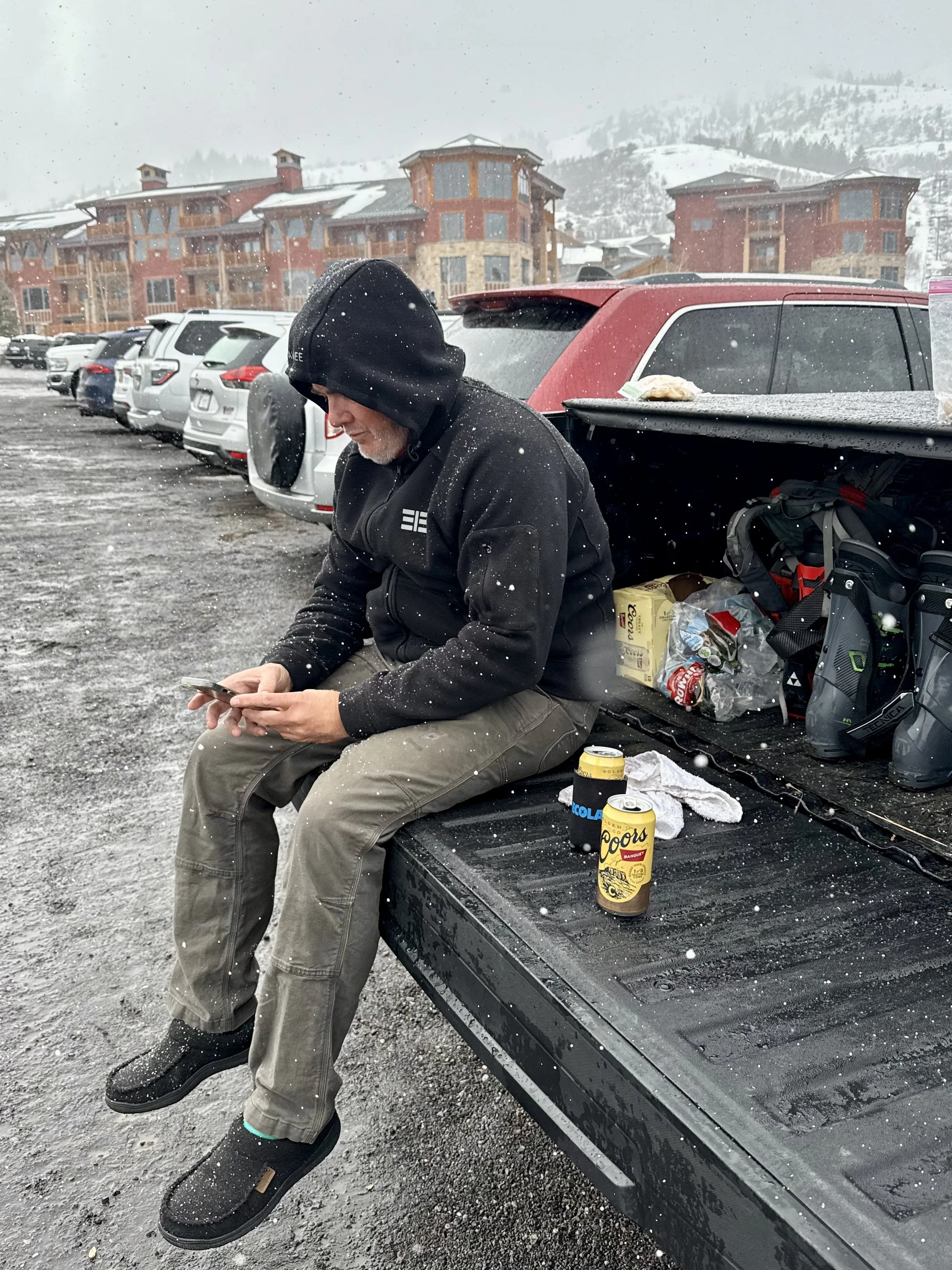 A man with a jacket sitting on the tailgate of a truck in a snowy parking lot. There are cans of Coors beer and snacks on the truck's tailgate. Multiple parked cars and a large apartment building are in the background.
