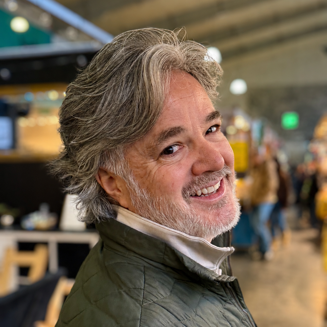 A cheerful older man with gray hair and a beard, smiling at the camera in a busy indoor setting.