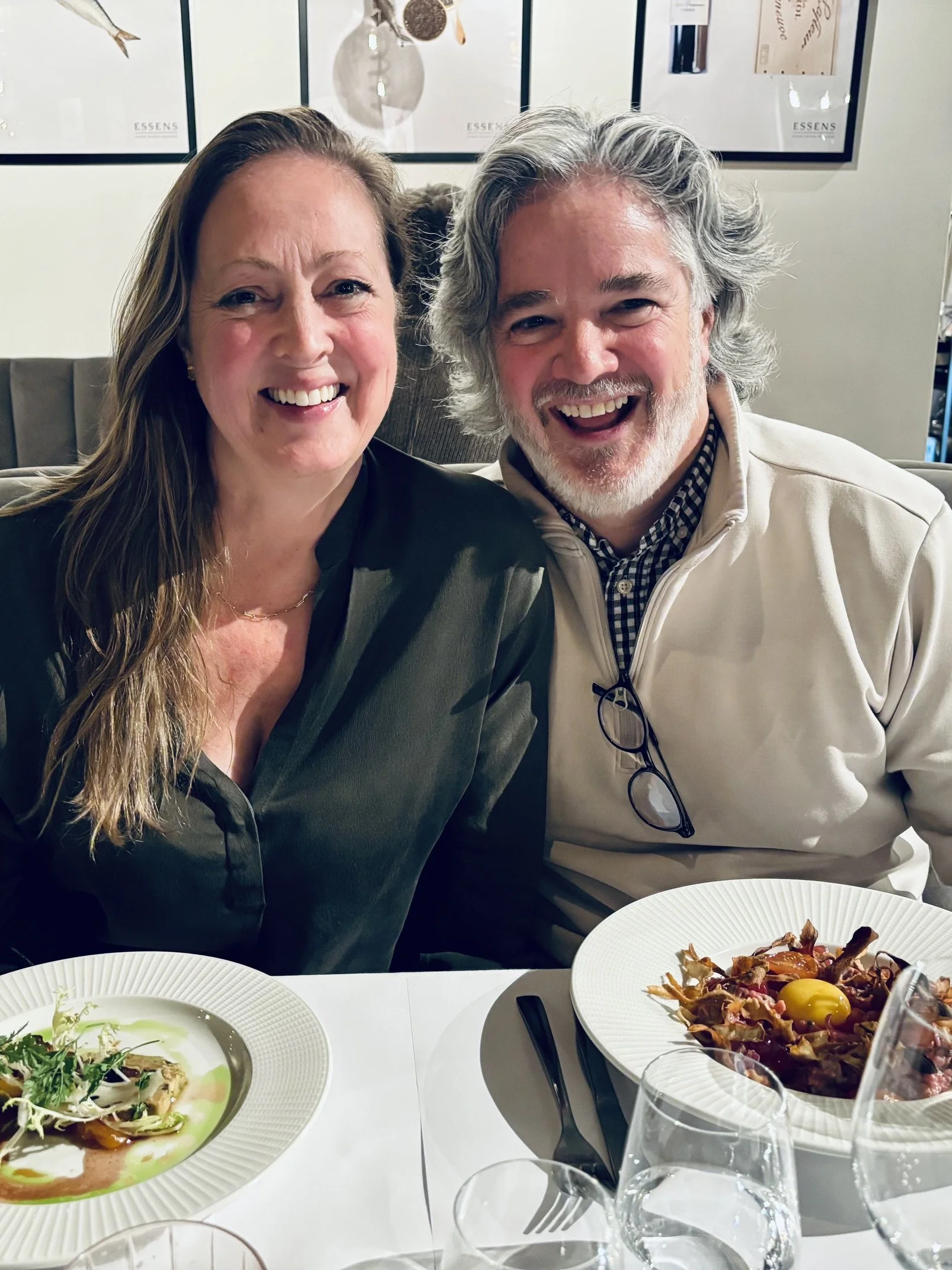 A smiling woman and man sitting together at a table in a restaurant with plates of food and glasses of water in front of them. The background shows framed pictures on the wall.