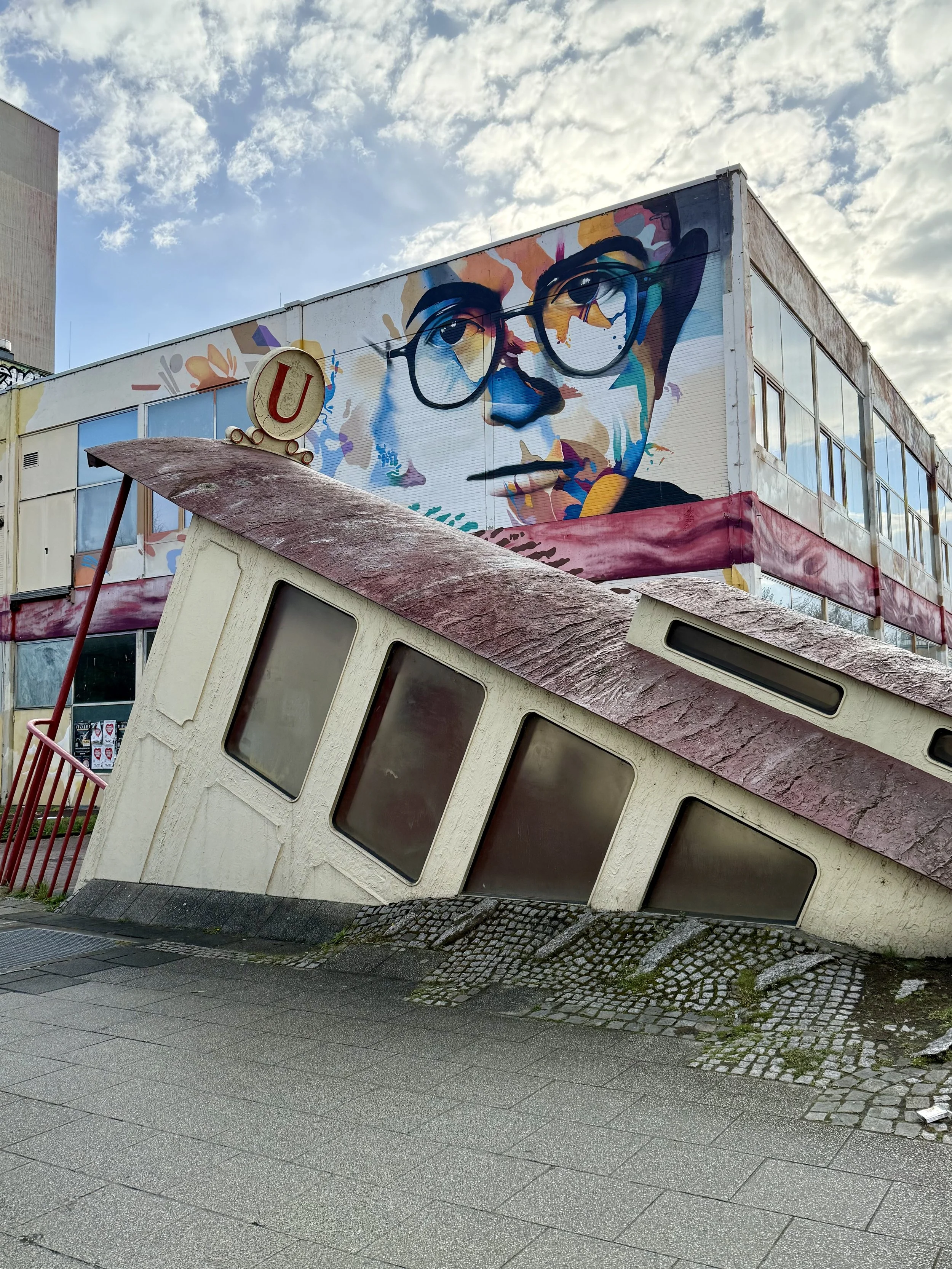 A colorful mural of a person wearing glasses painted on the side of a building. The building is slightly tilted in the image, with the foreground showing a patterned sidewalk and some toppled U-ban train entrance. The sky is partly cloudy.