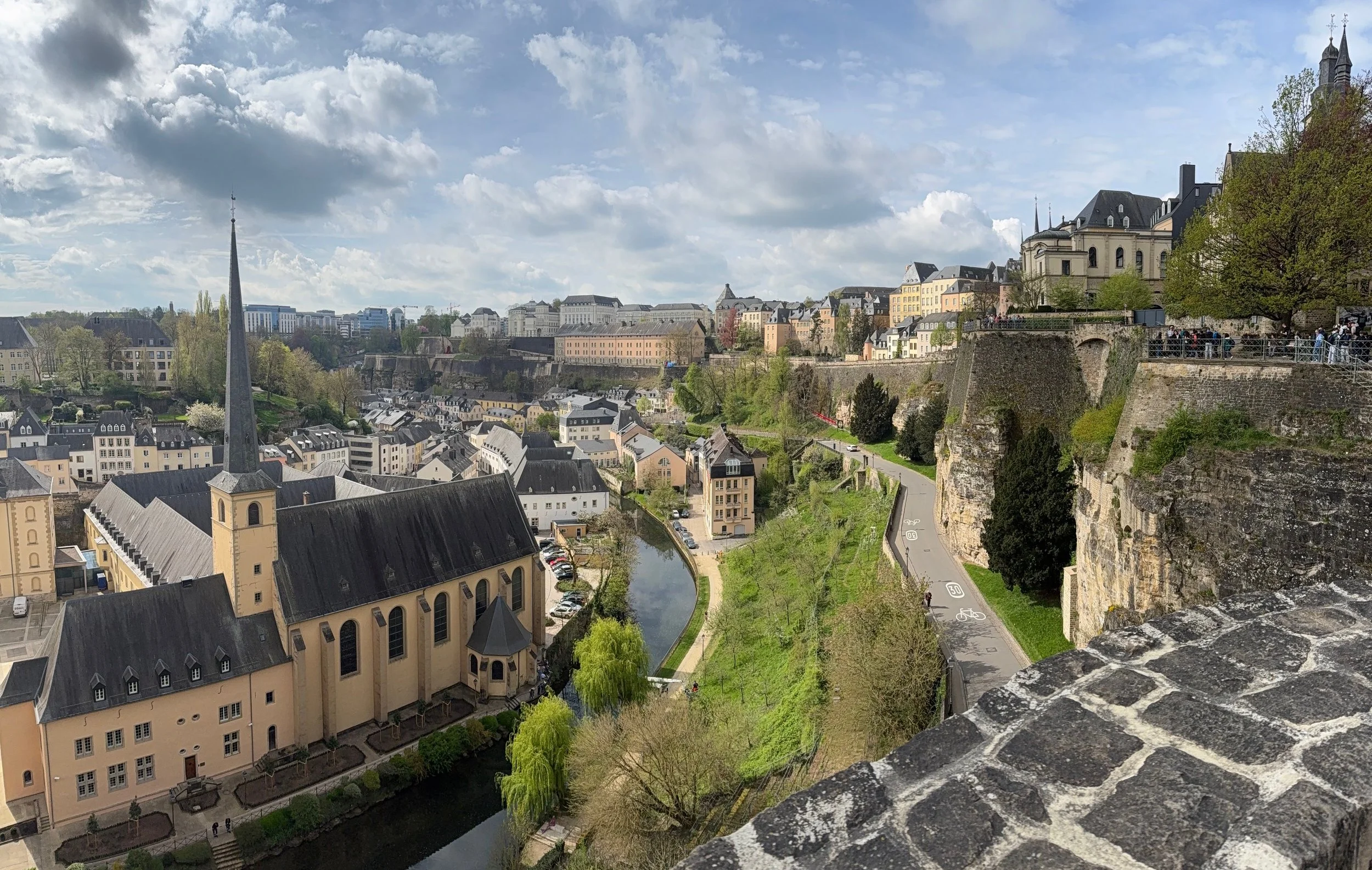 A panoramic view of Luxembourg City with a river, churches, and buildings on a hill under a cloudy sky.