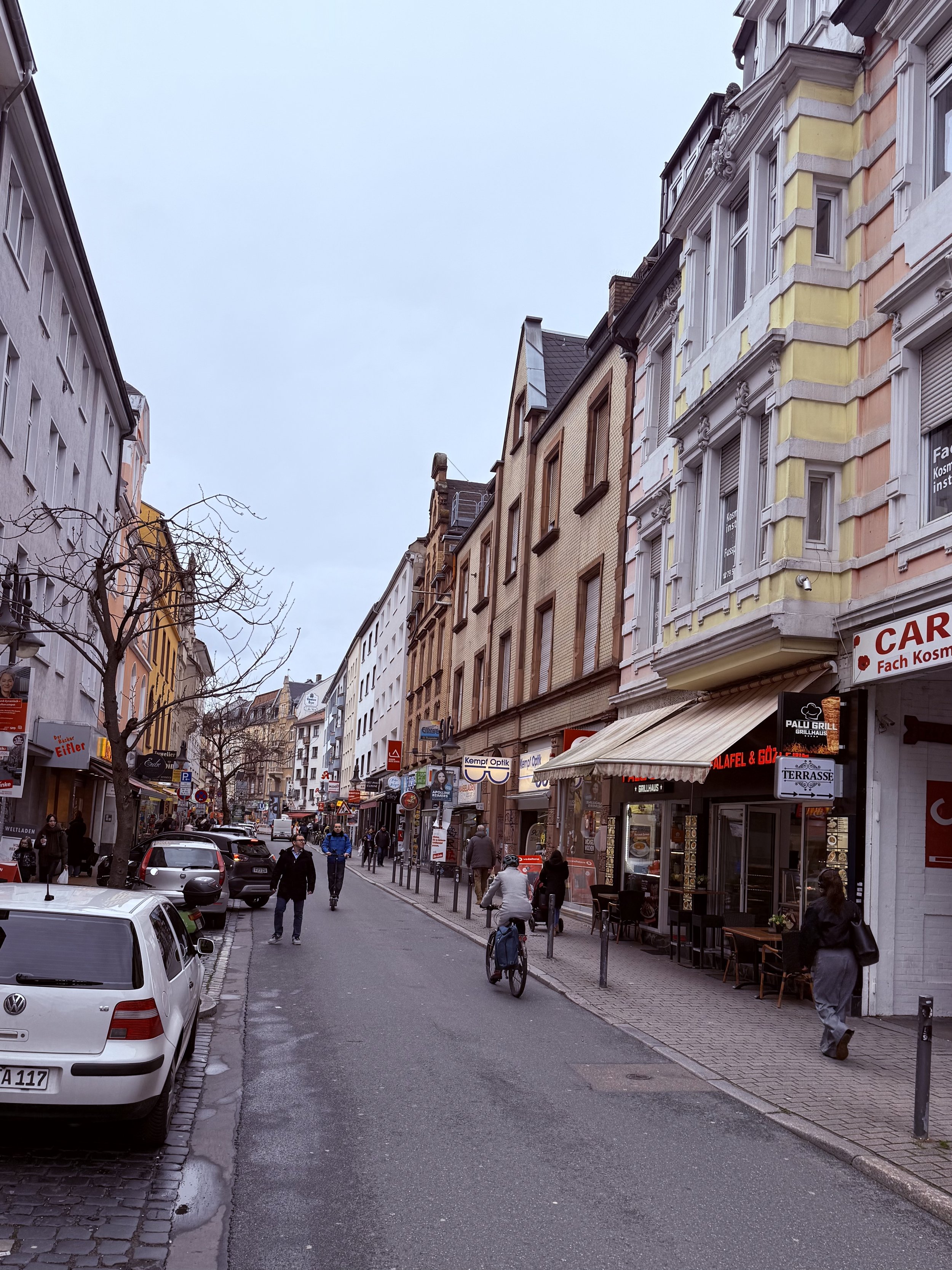 A street in the Bockenheim neighborhood of Frankfurt with cars parked along the side and people walking and riding bicycles. Buildings line both sides, with storefronts and signs visible, including a restaurant with outdoor seating.