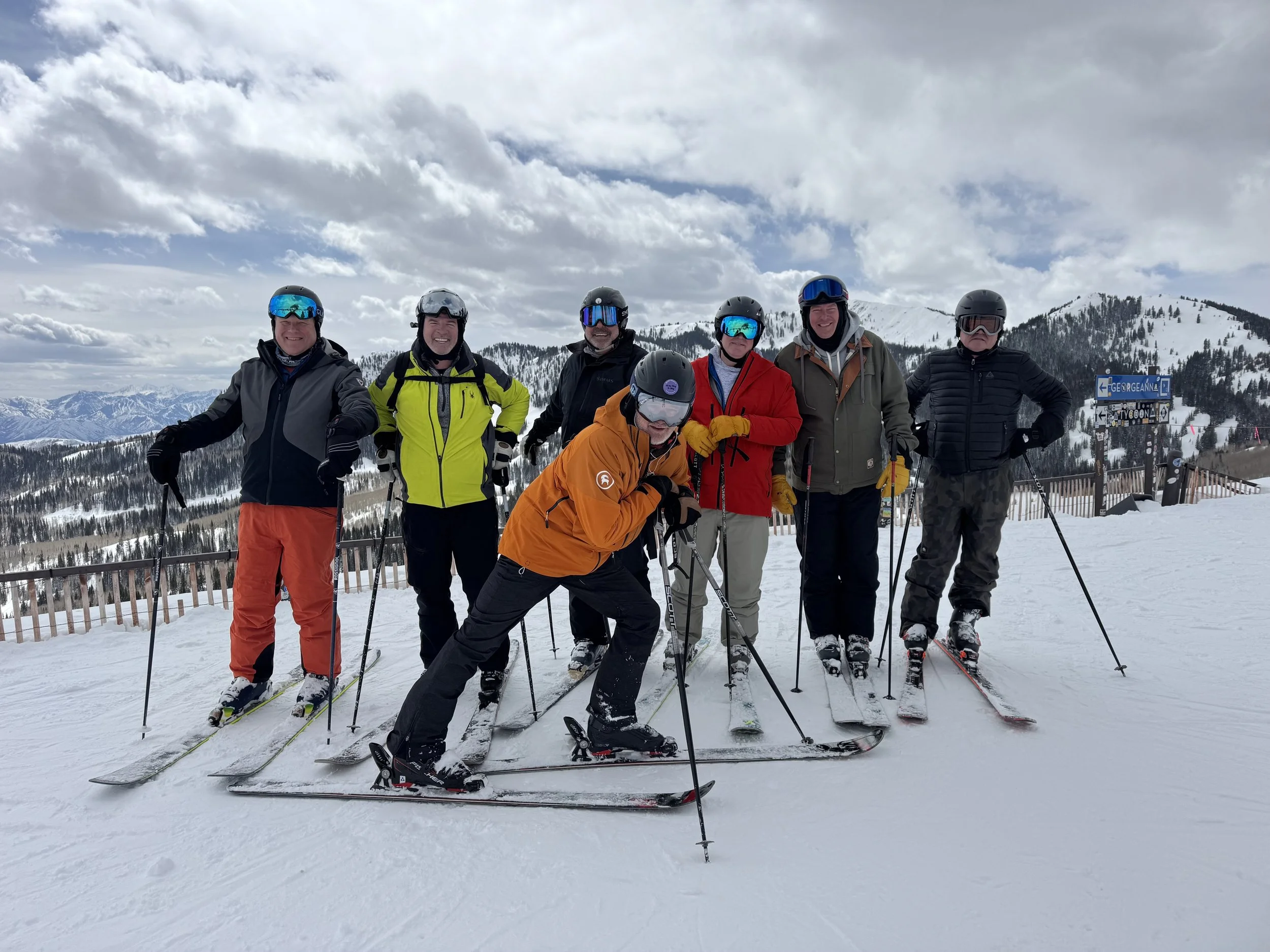 Group of seven people wearing ski gear and helmets, standing on a snowy Park City, Utah mountain with ski poles, smiling and posing for a photo. Snow-covered mountains and a cloudy sky in the background.