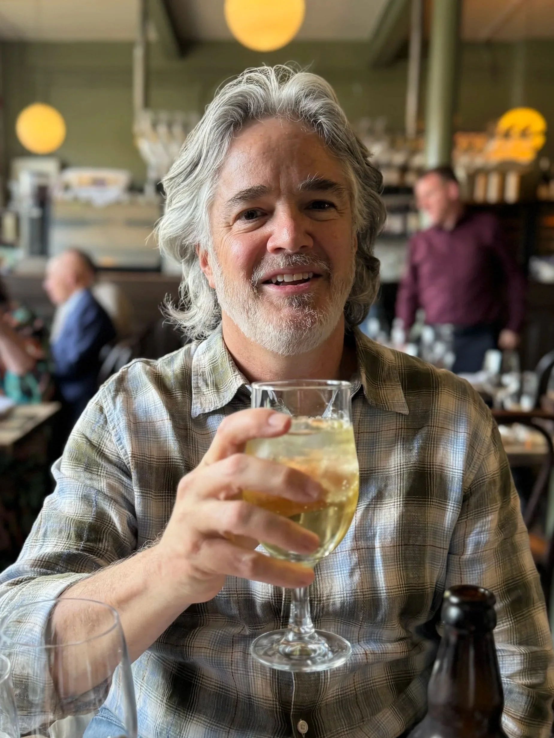 An older man with gray hair and a beard smiling and holding a glass of hard cider in a restaurant.