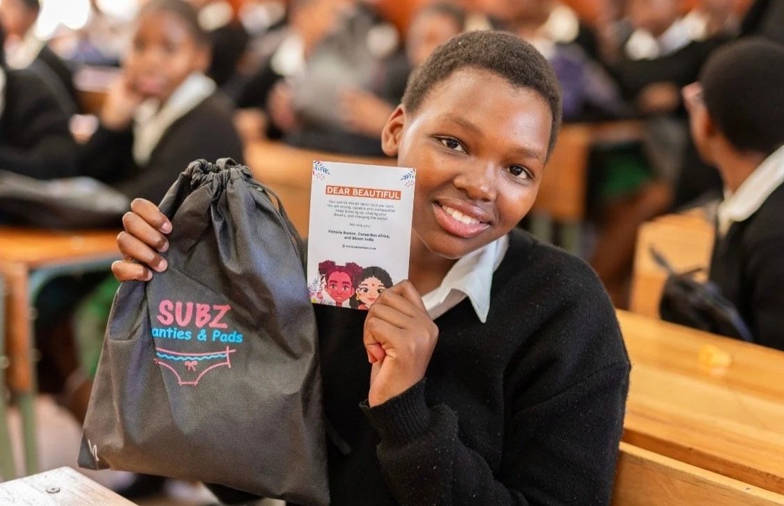 Schoolgirl smiling, holding a card and a drawstring bag labeled 'SUBZ Panties & Pads' in a classroom with other students in the background.