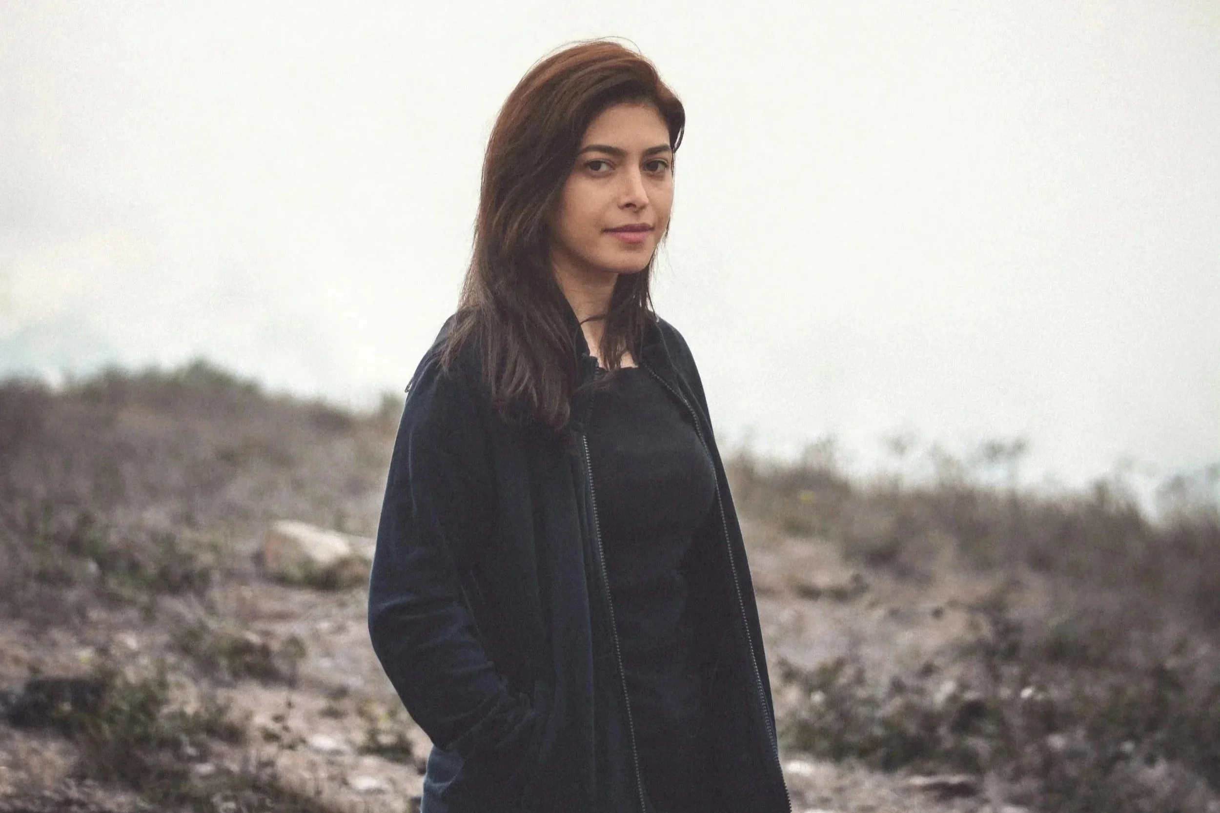 A woman with brown hair, wearing a black jacket, standing outdoors in a natural, overcast landscape.