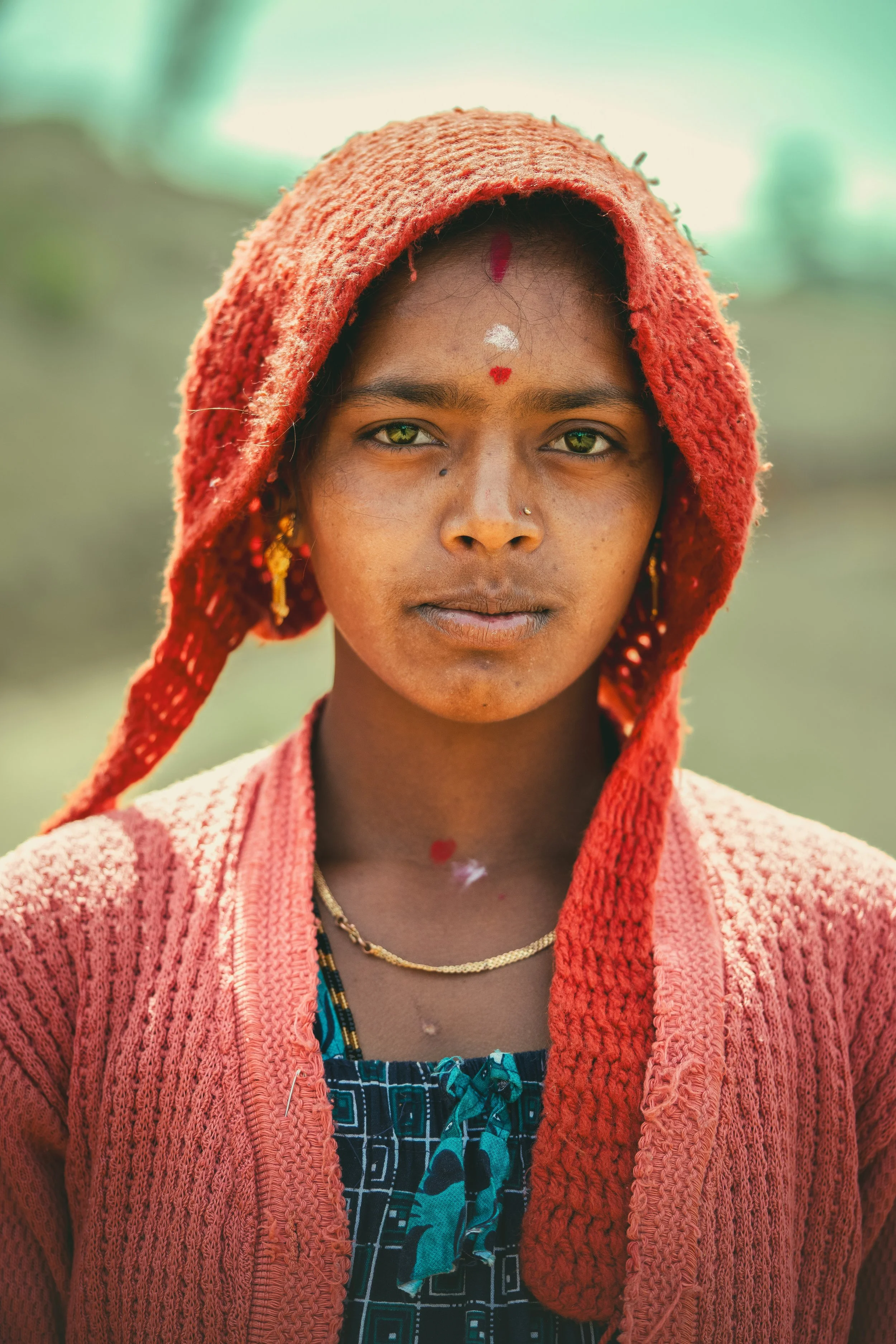 A woman with green eyes and traditional markings on her forehead and face, wearing a red hooded shawl, gold earrings, and a gold necklace, standing outdoors with a blurred green background.