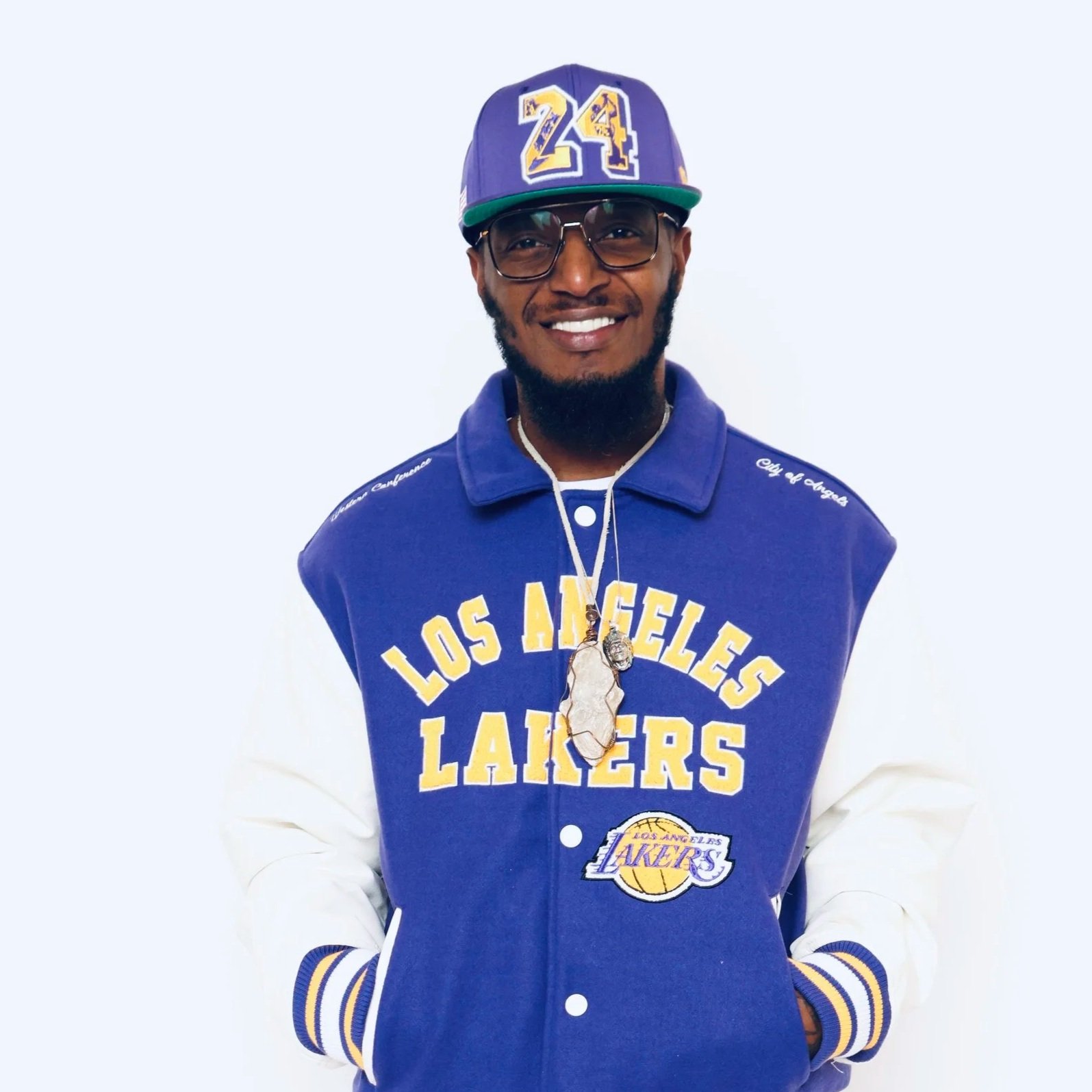 A man wearing a Los Angeles Lakers jacket, a cap with the number 24, glasses, and a necklace with a large pendant, smiling against a plain light background.