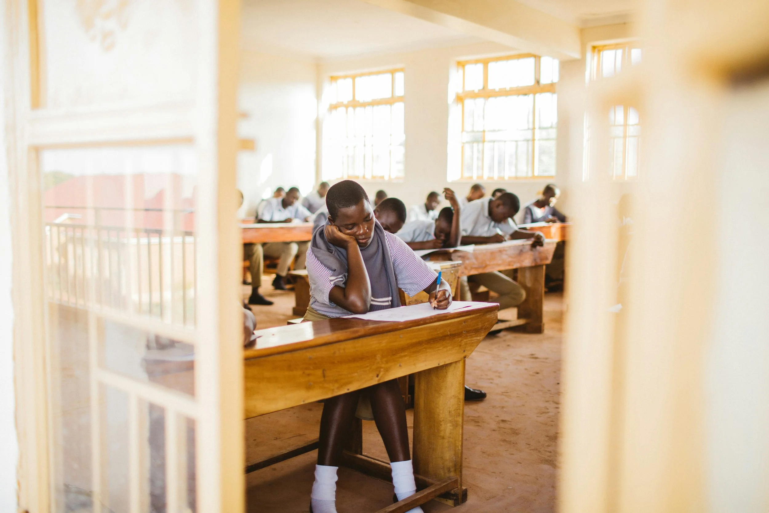 Students taking a test in a bright classroom, viewed through an open door with a girl in the foreground resting her head on her hand, writing on a paper.