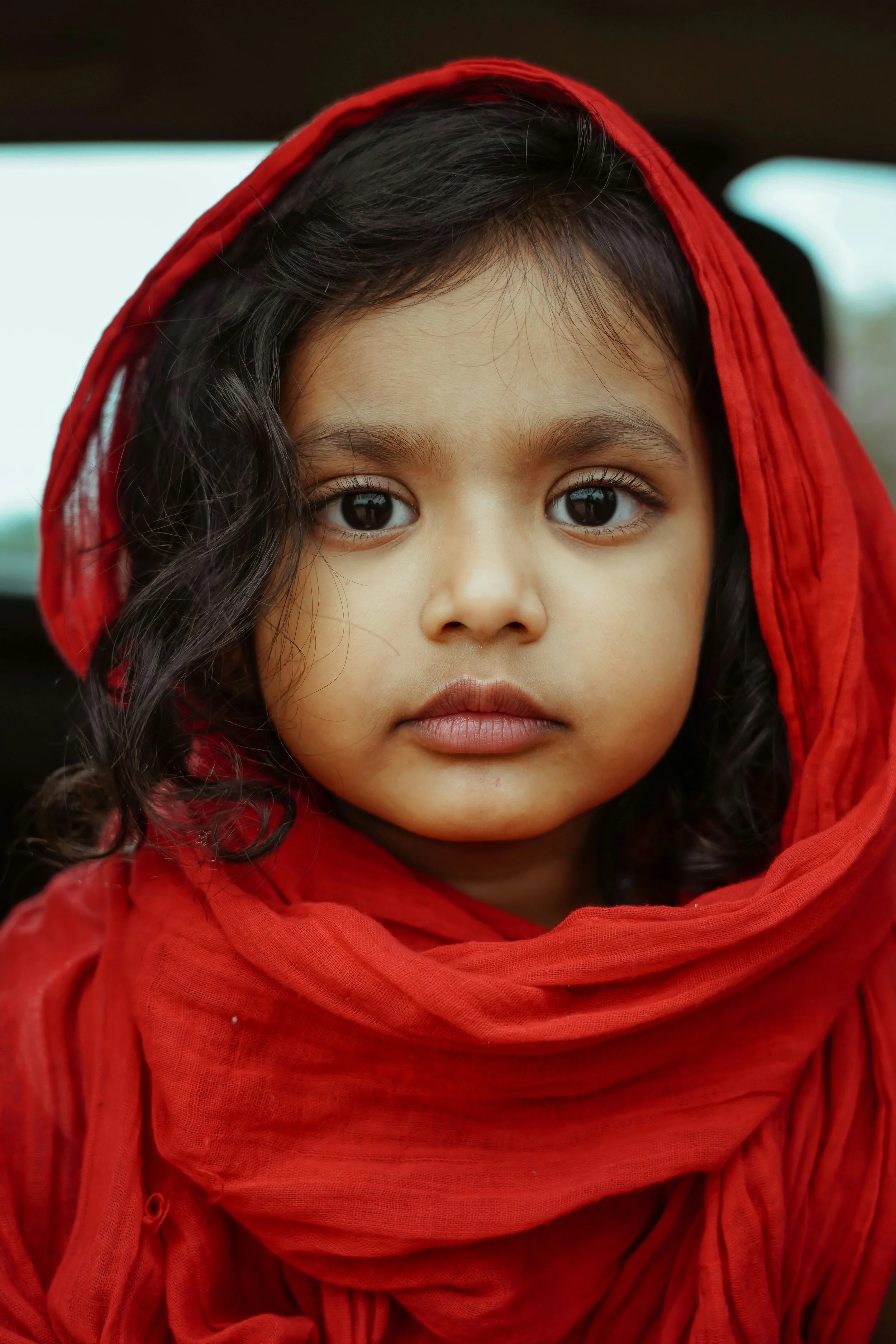A young girl wearing a red headscarf and red clothing, looking directly into the camera with a serious expression.