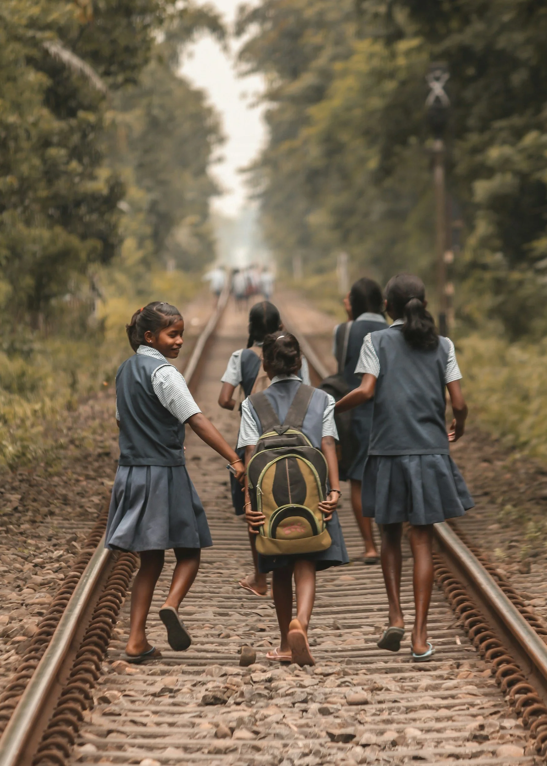 Group of young schoolgirls walking on railway tracks in a rural area, holding hands and carrying backpacks, with trees and power lines in the background.