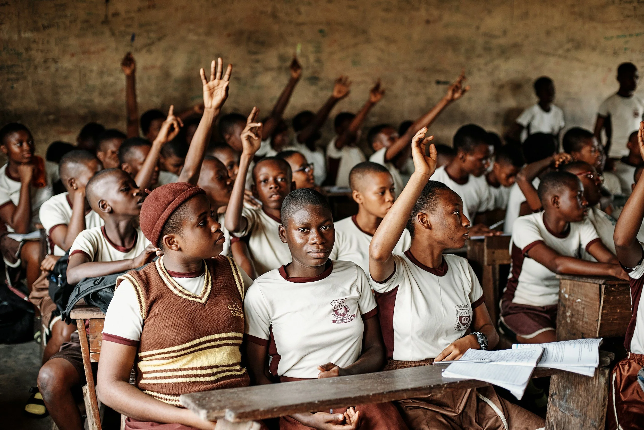 A classroom full of students with some raising their hands while others are sitting at desks, with a yellow and maroon uniform and notebooks.