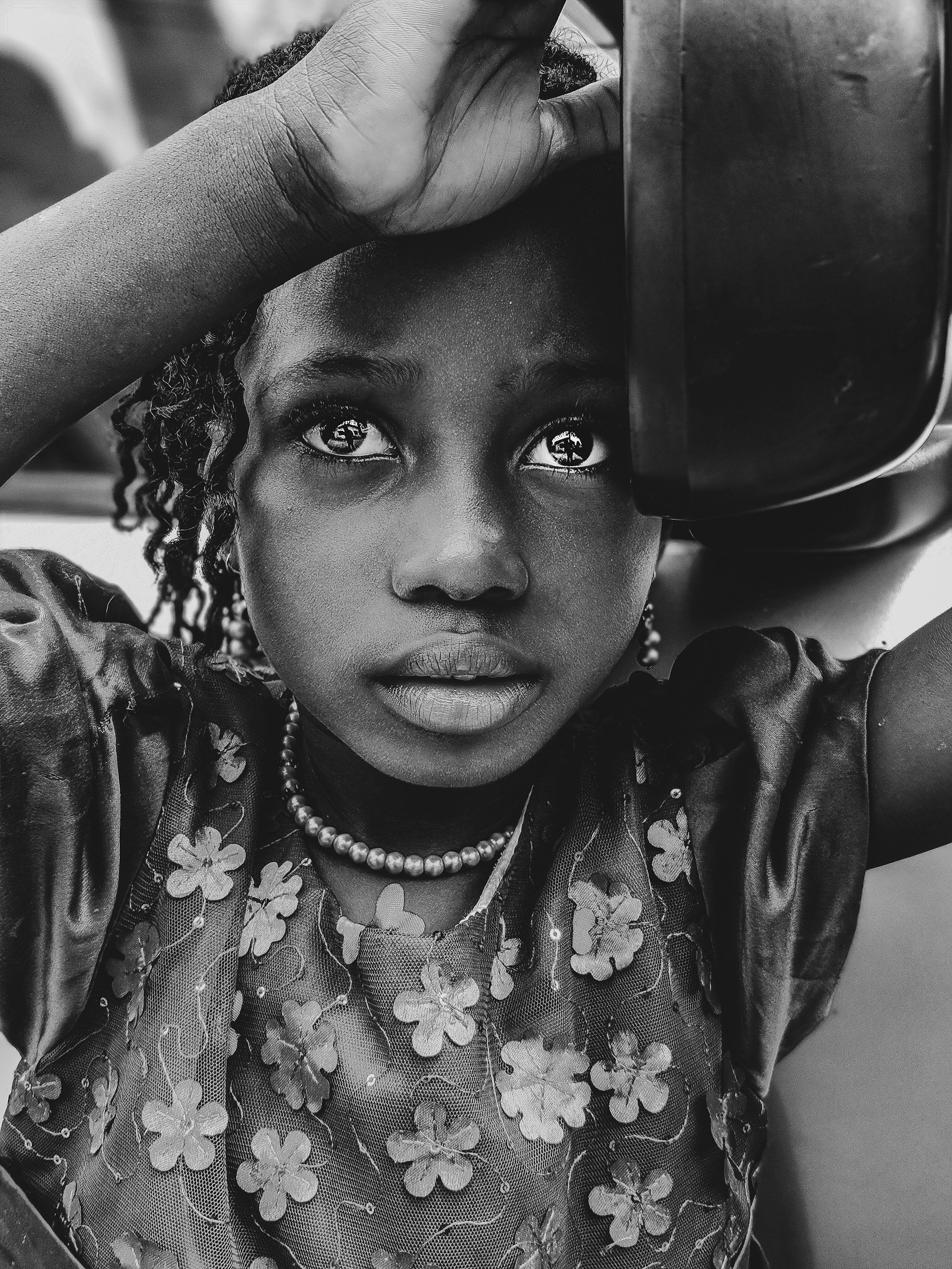 A black and white photograph of a woman looking into a mirror, wearing a floral-patterned top and a pearl necklace, with her hand resting on her forehead.