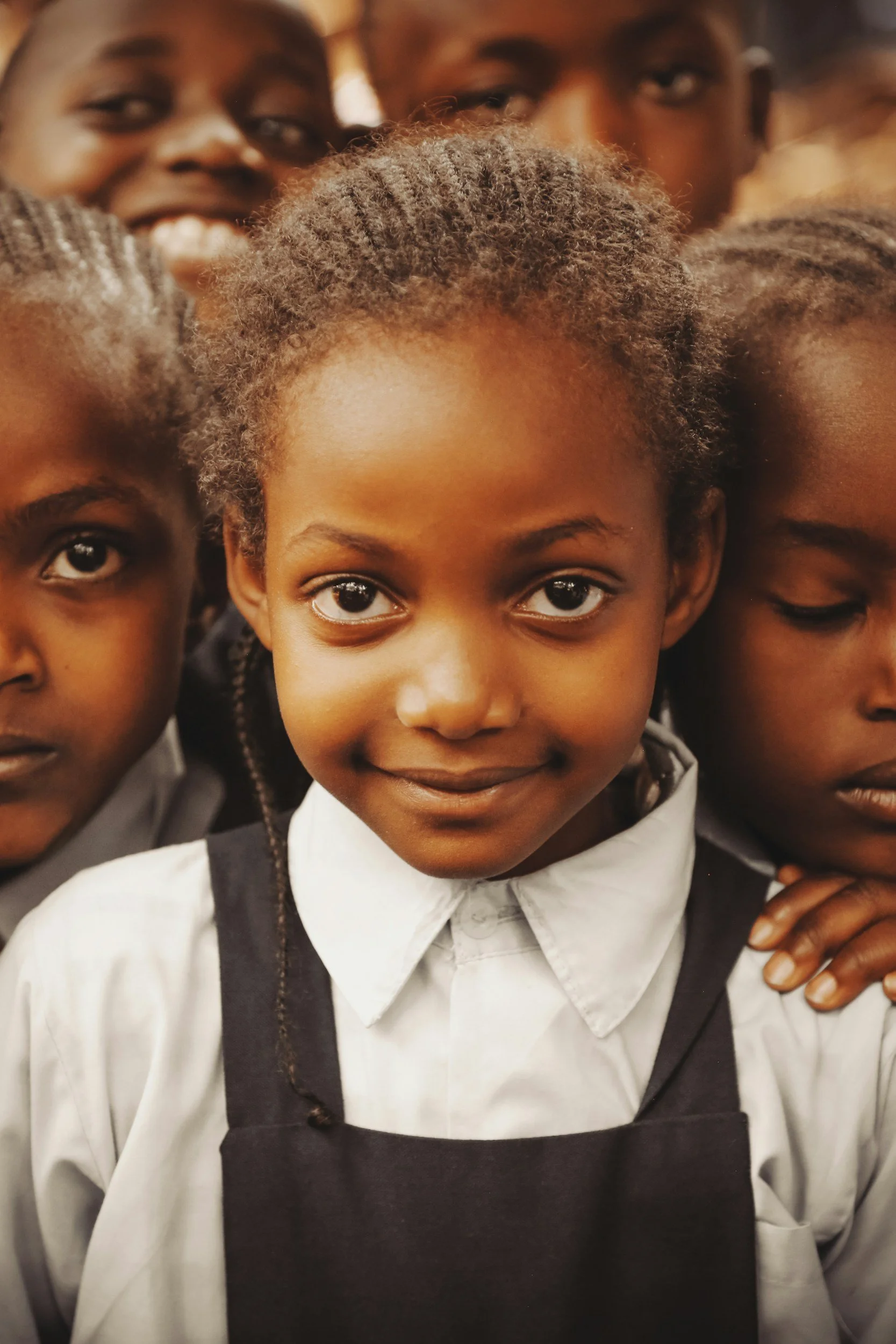 Close-up of a young girl with bright eyes and curly hair, wearing a white shirt and black apron, surrounded by other children in similar attire, smiling and looking at the camera.