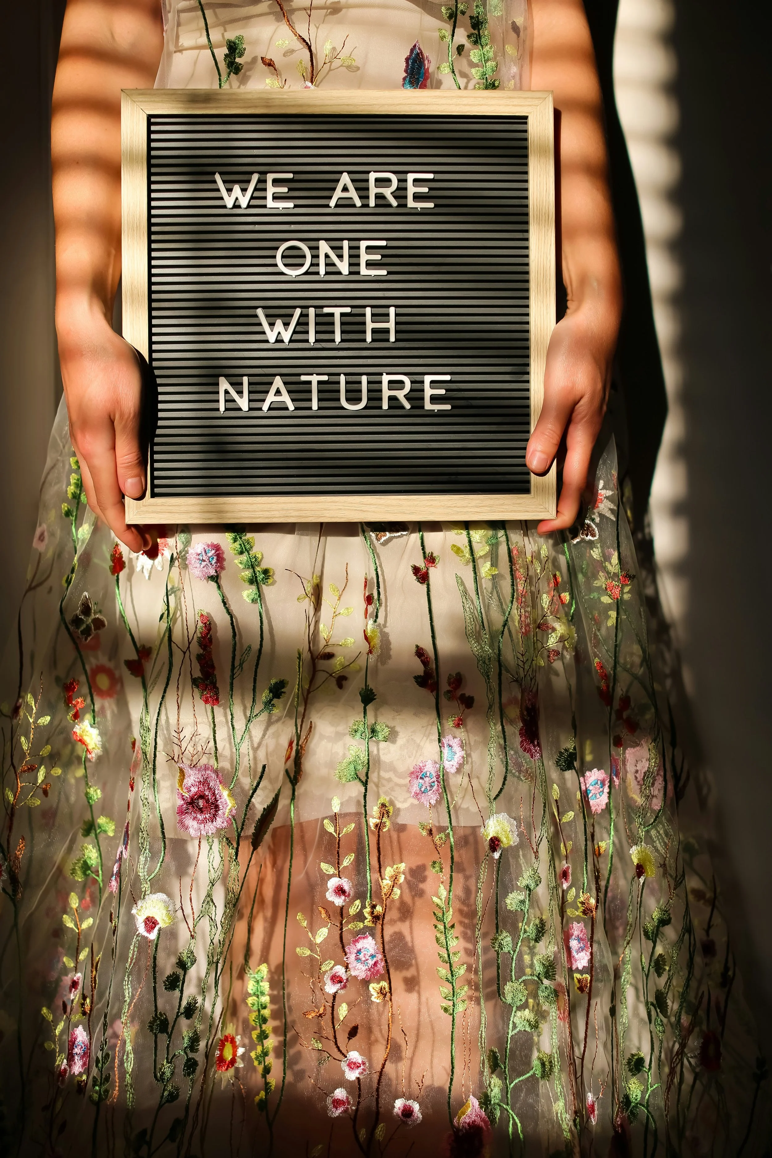 A person is holding a letter board that reads "WE ARE ONE WITH NATURE". The person is wearing a sheer, embroidered dress with floral patterns.