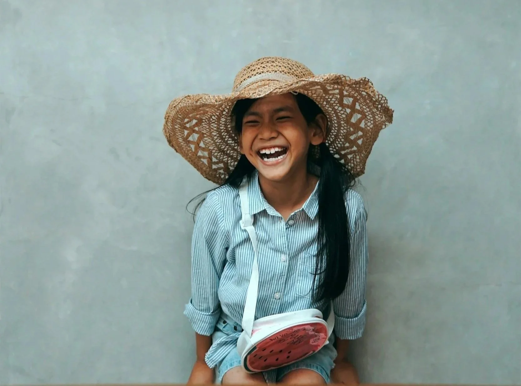A young girl with long black hair, smiling broadly and wearing a large straw hat and a light blue striped shirt, sitting against a plain light-colored wall. She is holding a bag designed to look like a watermelon.